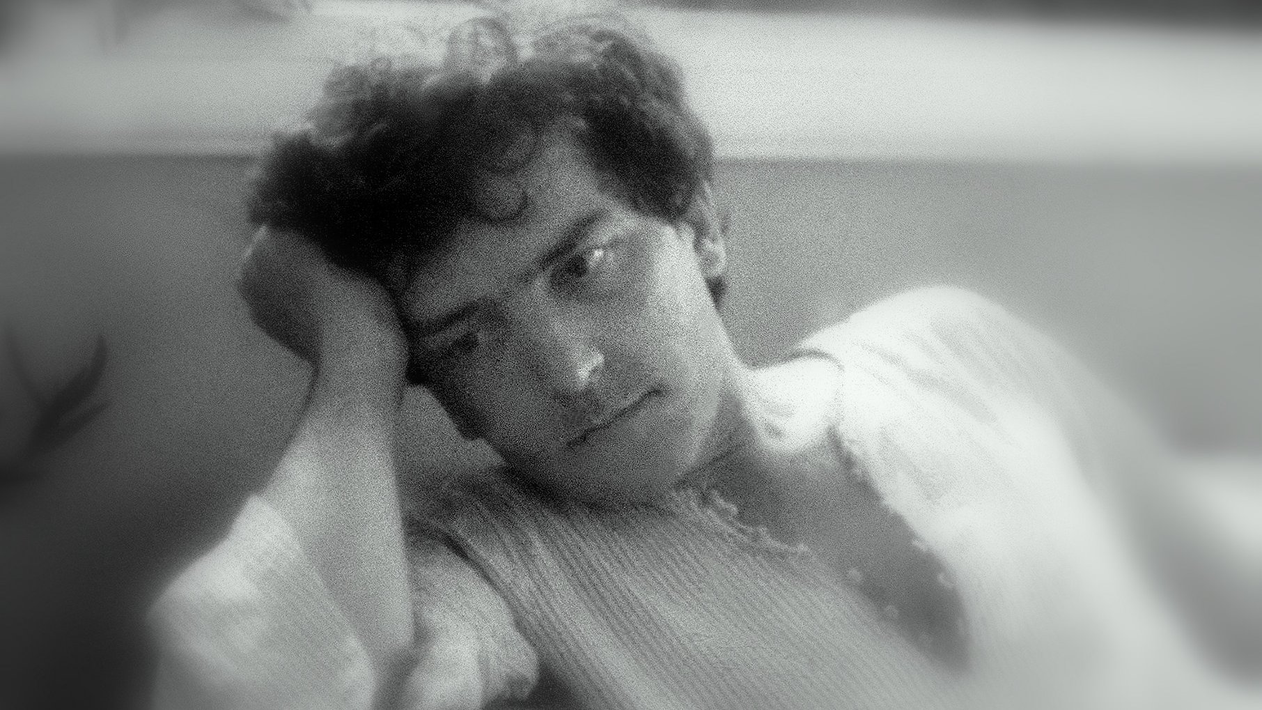 L'image montre un jeune homme pensif, appuyé sur sa main, avec une expression mélancolique. Il semble perdu dans ses pensées, ses cheveux sont légèrement ondulés et il porte une chemise à manches longues. L'arrière-plan est flou, créant une ambiance douce et introspective. La lumière est tamisée, ajoutant à l'atmosphère de réflexion.