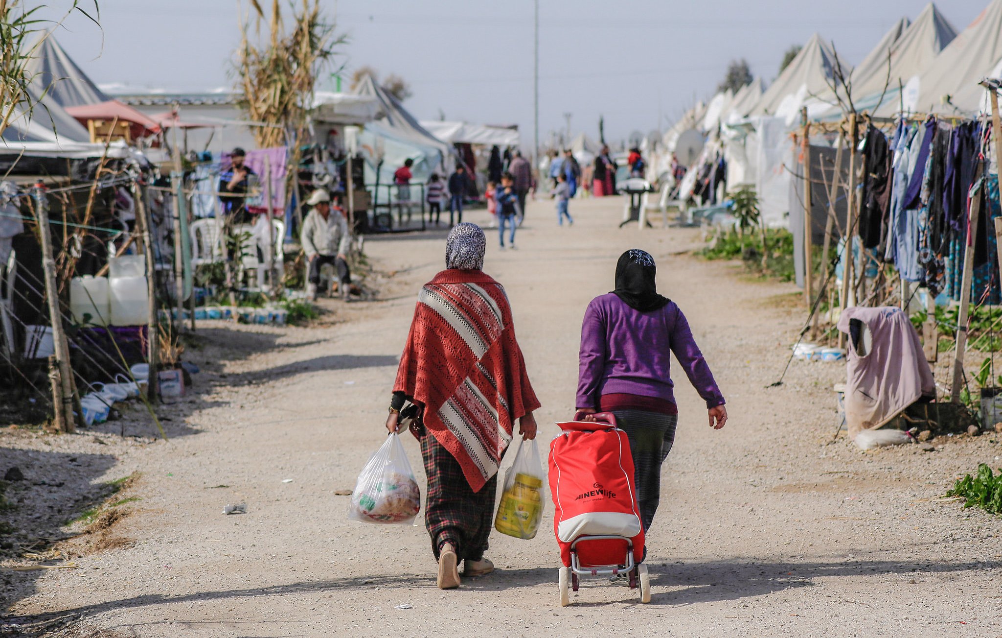 L'image montre un camp de réfugiés ou un espace similaire, avec plusieurs tentes dressées de chaque côté d'un chemin central. Deux femmes marchent le long de ce chemin, portant des sacs et un chariot. L'une d'elles porte une couverture ou un châle coloré. En arrière-plan, on peut apercevoir d'autres personnes et des tentes, suggérant une atmosphère de communauté. Le paysage semble rural et ensoleillé, avec des éléments de végétation autour.