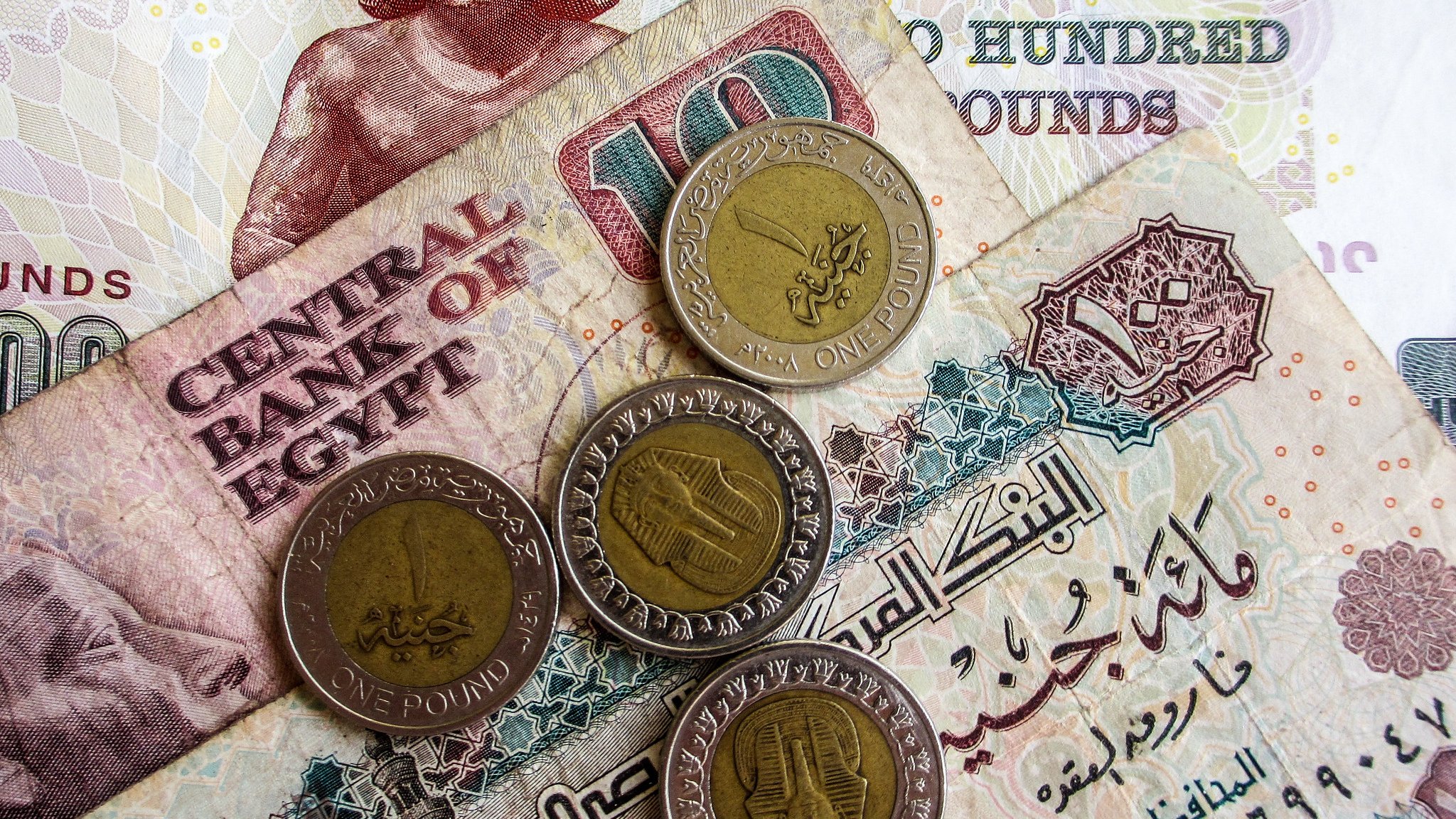 تظهر في الصورة مجموعة من العملات المصرية. هناك ورقة نقدية من فئة المائة جنيه بالإضافة إلى فئة العشرة جنيه، مع عدد من العملات المعدنية من فئة الجنيه الواحد. التصميمات المختلفة توضح رموز ومعالم ثقافية مرتبطة بمصر، مما يعكس التنوع في العملة المصرية.