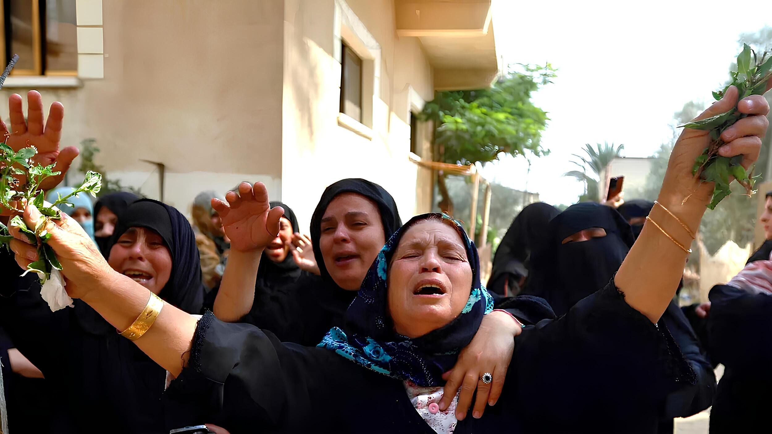 L'immagine mostra un gruppo di donne in un contesto emotivo intenso. Indossano abiti scuri e alcune di loro hanno il viso coperto. Alcune donne sembrano piangere e sollevano le braccia, mentre altre mostrano espressioni di sofferenza. In mano hanno foglie verdi, che possono rappresentare un simbolo di lutto o commemorazione. Sullo sfondo, si intravedono edifici e vegetazione. L'atmosfera è carica di emotività e partecipazione.