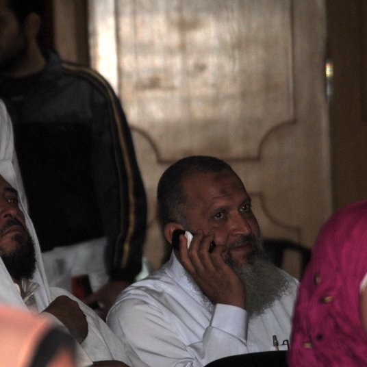 L'image montre des hommes et des femmes dans un environnement qui semble être un rassemblement ou une conférence. Deux hommes au premier plan portent des vêtements traditionnels, l'un d'eux est exprimé en train de passer un coup de téléphone. L'arrière-plan montre d'autres personnes, certaines concentrées sur l'événement, et des couleurs variées dans les vêtements, y compris une femme portant un hijab rose. L'atmosphère semble sérieuse et attentive.
