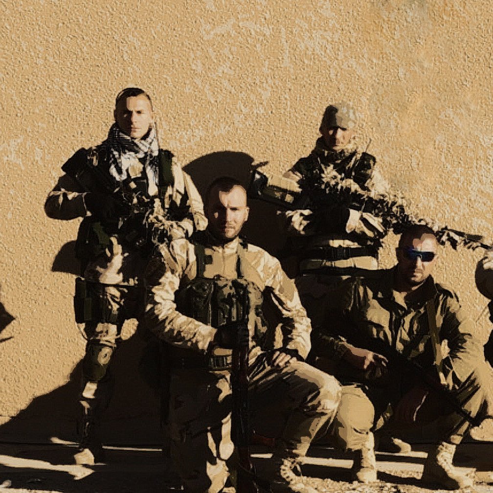 L'image montre un groupe de soldats en tenue militaire. Ils sont positionnés à la fois debout et accroupis, avec des armes à feu en main. L'arrière-plan est une surface murale de couleur beige, ce qui suggère un environnement désertique. Les soldats portent des uniformes camouflés et semblent prêts pour une mission. L'éclairage est plutôt clair, mettant en valeur leurs expressions et leurs équipements.