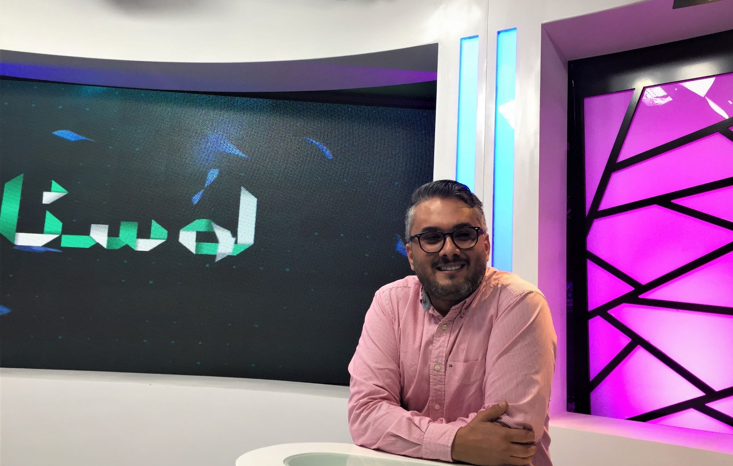 L'image montre un homme souriant assis devant un bureau dans un studio de télévision. Il porte une chemise rose et semble détendu. En arrière-plan, on peut voir un écran avec un motif abstrait et des lumières qui donnent une ambiance moderne au lieu. Le mot "لواح" est affiché sur l'écran, indiquant peut-être le titre d'une émission ou d'une rubrique. L'éclairage du studio est bien conçu, créant une atmosphère conviviale.