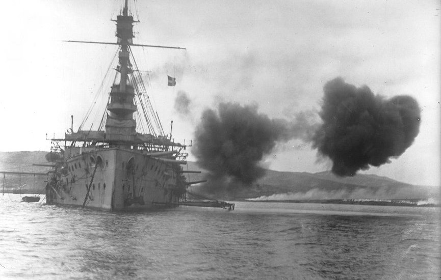 L'image montre un grand navire de guerre, probablement un cuirassé, tirant des armes à feu, ce qui crée des nuages de fumée sur l'eau. Le navire est équipé de plusieurs canons sur ses flancs. En arrière-plan, on aperçoit des collines ou des montagnes, ainsi qu'une éventuelle base maritime ou un autre type d'installations côtières. L'atmosphère est dynamique, accentuée par l'action de tir.