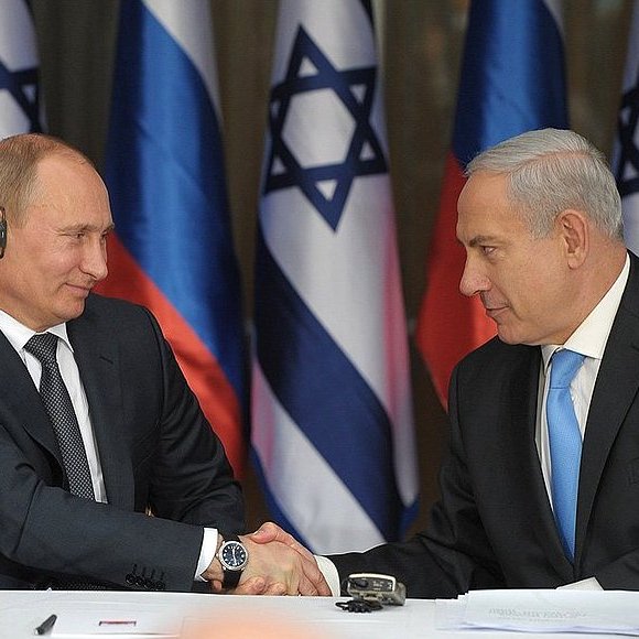 تظهر الصورة اجتماعاً رسميًا بين شخصين. يجلس أحدهما على اليسار، يرتدي بدلة داكنة مع سماعة أذن، في حين يجلس الآخر على اليمين ويبدو أنه يرتدي بدلة فاتحة مع رابطة عنق زرقاء. كلا الشخصين يتصافحان فيما يبدو كعلامة على الاتفاق أو التعاون، مع وجود خلفية تحتوي على أعلام متعددة.