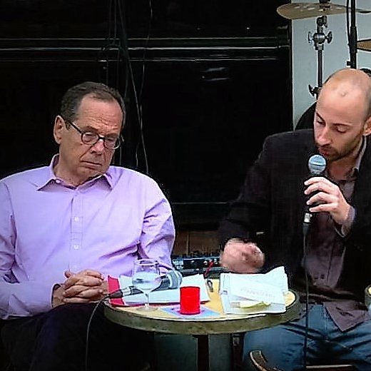 L'image montre un moment lors d'un événement où trois hommes sont assis autour de petites tables. Au centre, un homme parle dans un micro, semblant engager une discussion ou une présentation. Les autres hommes, à sa gauche et à sa droite, écoutent attentivement. On peut voir des instruments de musique en arrière-plan, ce qui suggère que l'événement se déroule dans un cadre décontracté, peut-être un café ou un bar. Des verres et des notes sont présents sur les tables, ajoutant à l'ambiance informelle de la rencontre.