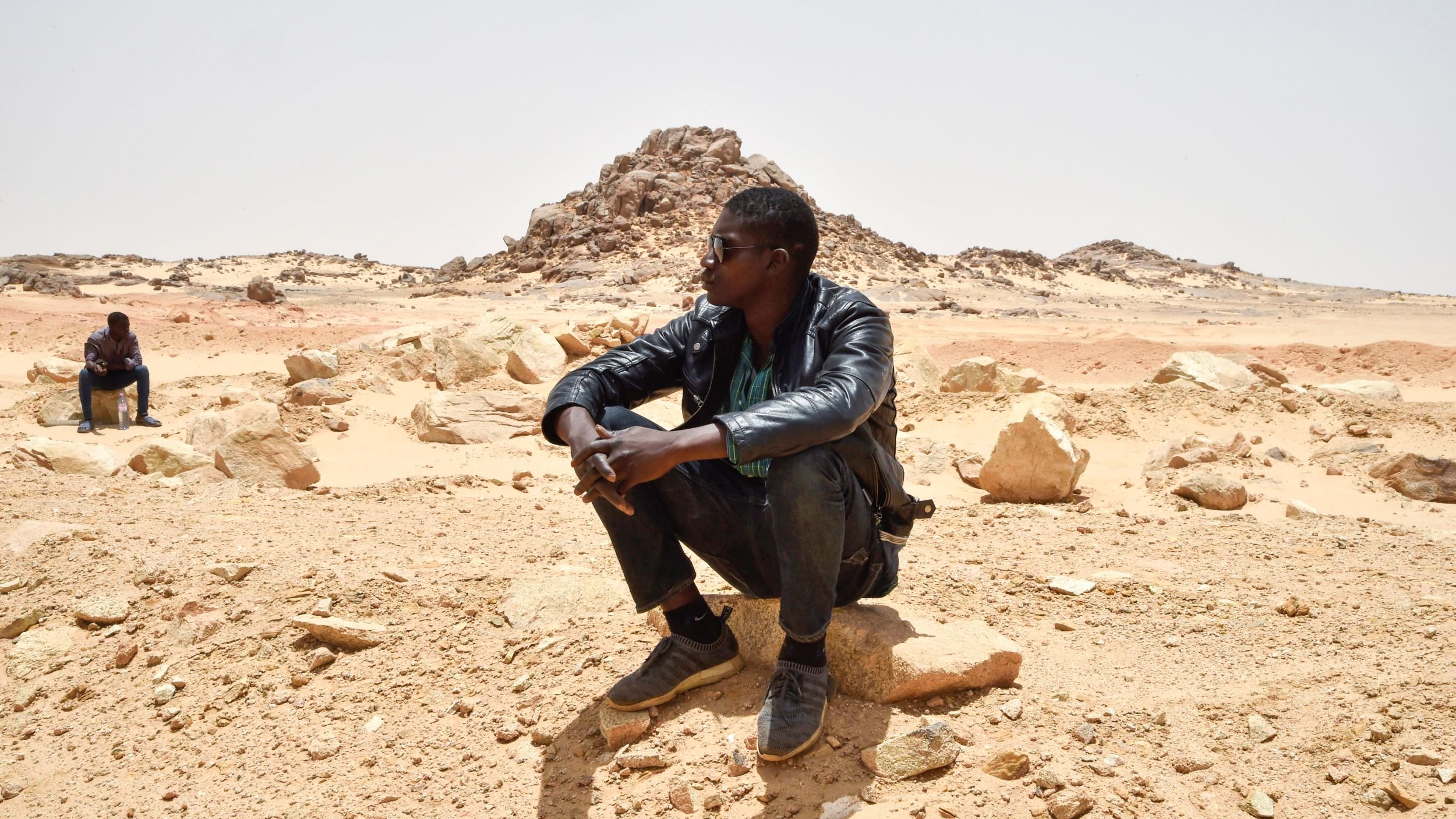 تظهر الصورة رجلين يجلسان في منطقة صحراوية. أحدهما يجلس على صخرة ويبدو عليه التفكير، بينما الآخر يجلس في الخلف بعيدًا قليلاً. الخلفية تتكون من صخور وكثبان رملية تحت سماء صافية. يبدو المشهد هادئًا، مما يعكس طبيعة الصحراء القاحلة.