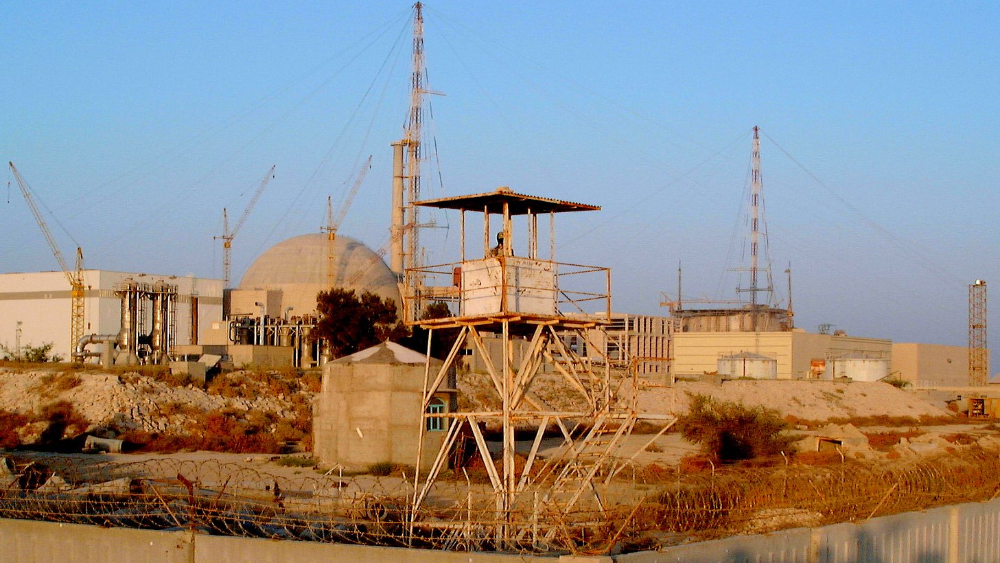 L'image présente un site industriel, probablement une centrale nucléaire, avec plusieurs structures. On peut voir une grande infrastructure circulaire au fond, entourée de diverses installations et grue. Au premier plan, il y a une tour de garde sur une plateforme surélevée, indiquant probablement un niveau de sécurité élevé dans cette zone. Des barbelés et des éléments de sécurité entourent l'endroit, suggérant une zone protégée. Le ciel est clair, ce qui donne une lumière naturelle à la scène.