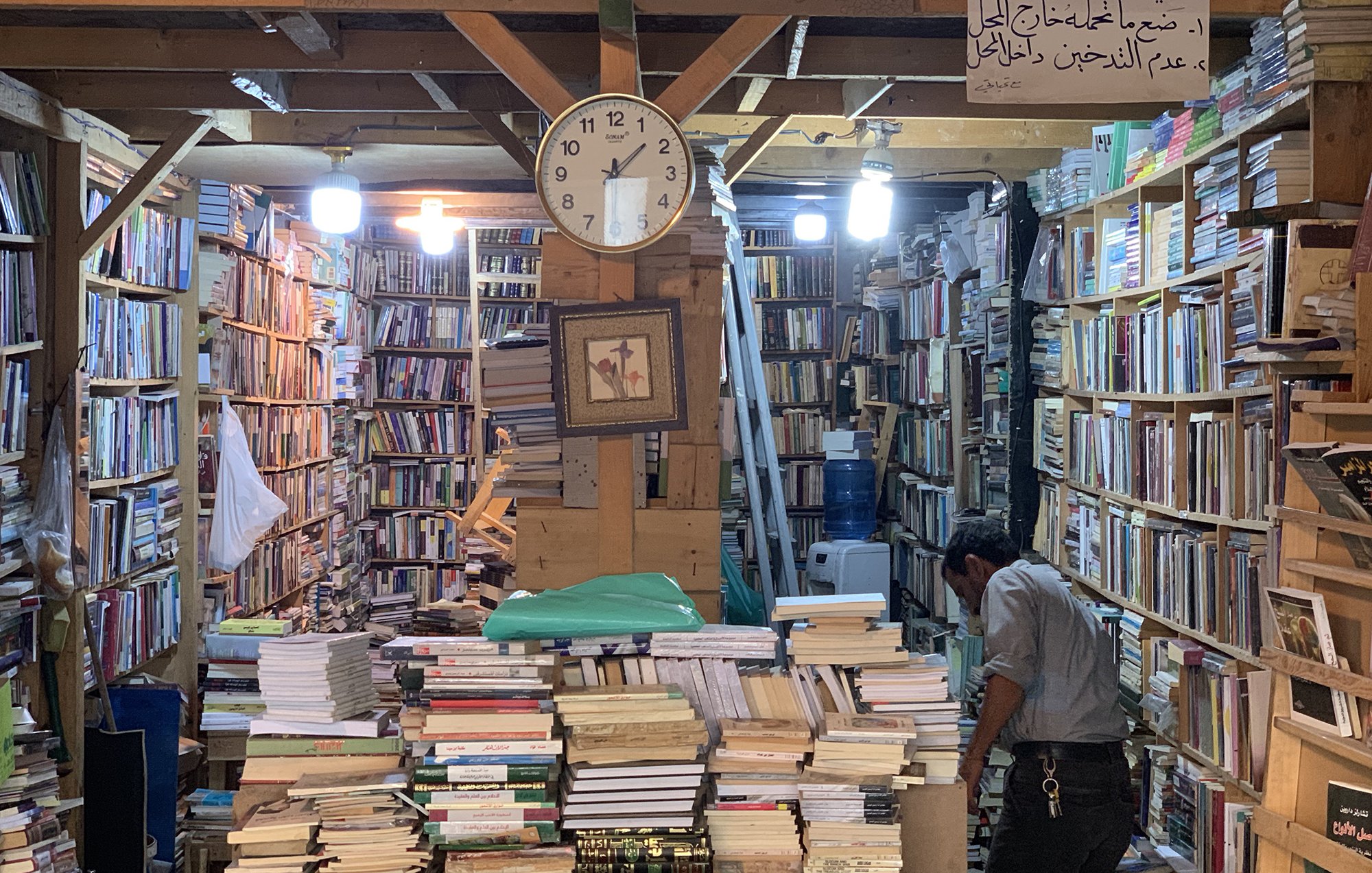 L'image montre une librairie pleine de livres, avec des étagères qui montent jusqu'au plafond. Il y a une grande horloge murale qui indique l'heure. Au centre, des piles de livres sont empilées sur une table, tandis qu'un homme est penché sur une étagère, en train de chercher ou de réorganiser des livres. L'ambiance semble chaleureuse et accueillante, avec un éclairage doux provenant de lampes suspendues. Une inscription en arabe est visible au mur.