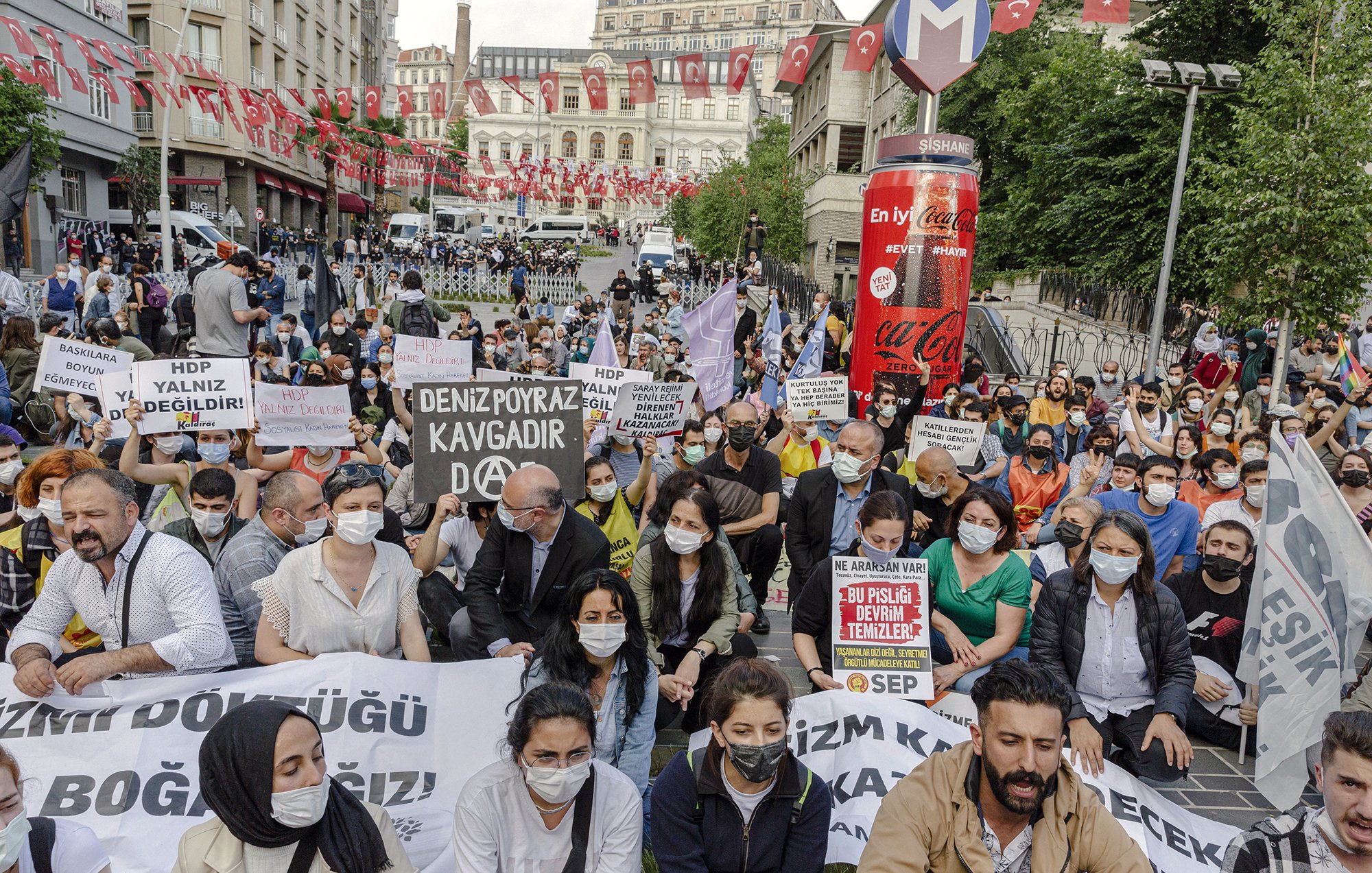 La imagen muestra una manifestación en una plaza urbana, con varias personas sentadas en el suelo. Los participantes sostienen pancartas y carteles que tienen mensajes de protesta. La mayoría de las personas llevan mascarillas, lo que puede indicar que la manifestación está ocurriendo en un contexto reciente de pandemia. Al fondo, se pueden ver banderas turcas y edificios, lo que sugiere que la protesta tiene lugar en Turquía. La atmósfera es de movilización y reclamo social.