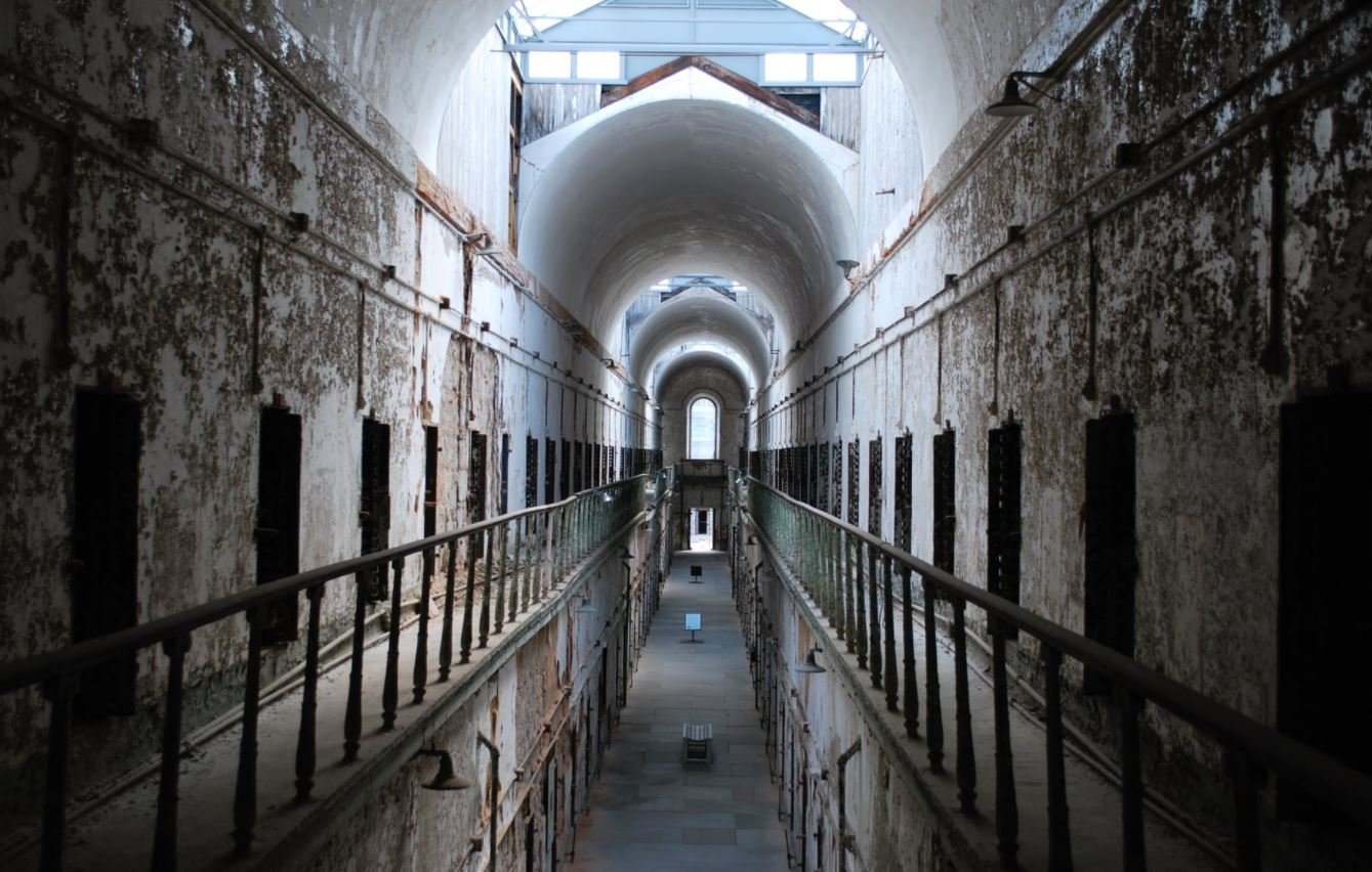 L'image montre l'intérieur d'une ancienne prison, avec un long couloir central bordé de cellules. Les murs sont décolorés et décrépis, témoignant de l'âge de l'établissement. Le plafond est haut, avec de grandes arches et une lumière qui pénètre par des lucarnes au-dessus. Les balustrades en fer forgé entourent les niveaux supérieurs, ajoutant à la structure vétuste. L'ambiance générale suggère une atmosphère d'abandon et d'histoire.