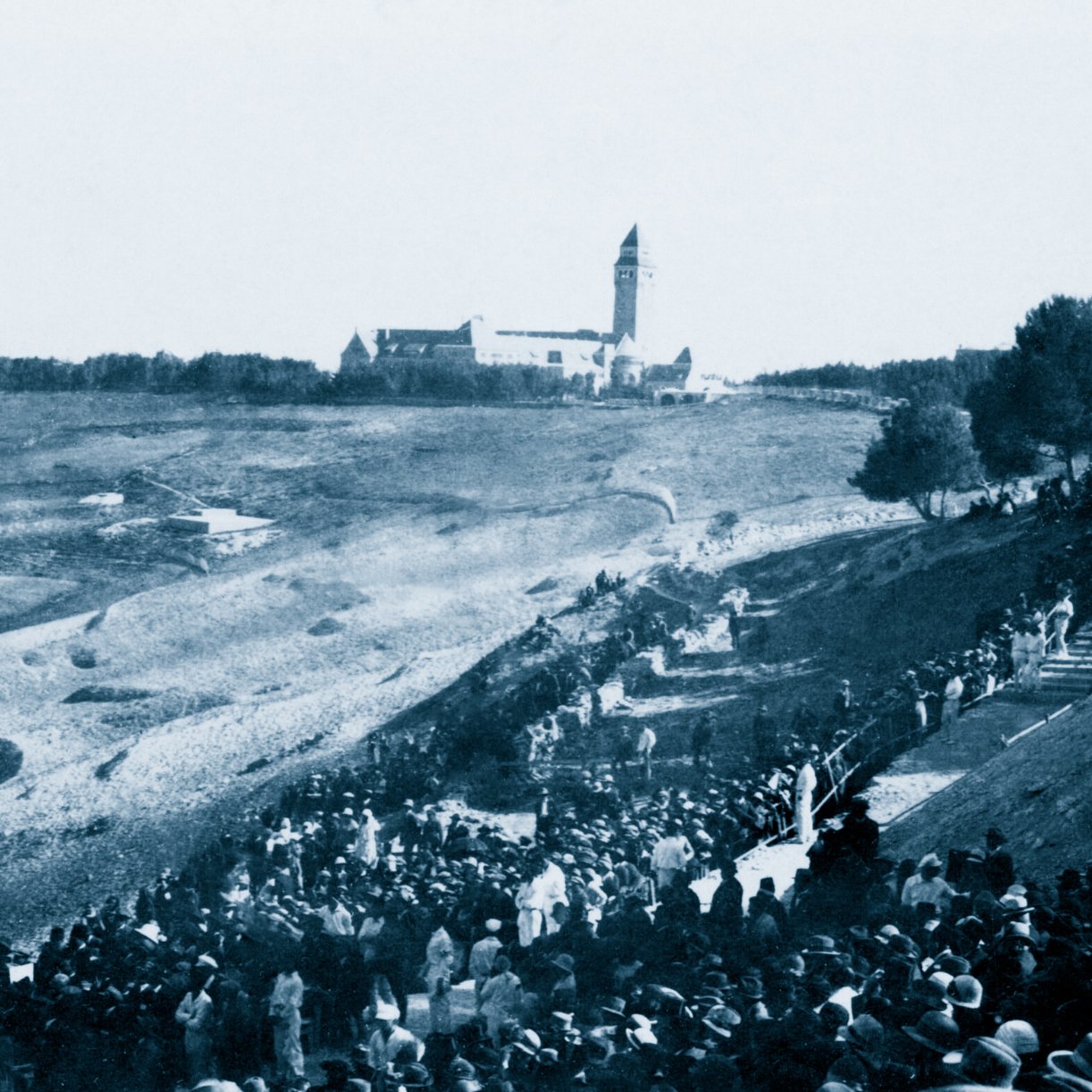 A large crowd in historical clothing gathers on a hillside, with a building in the distance.