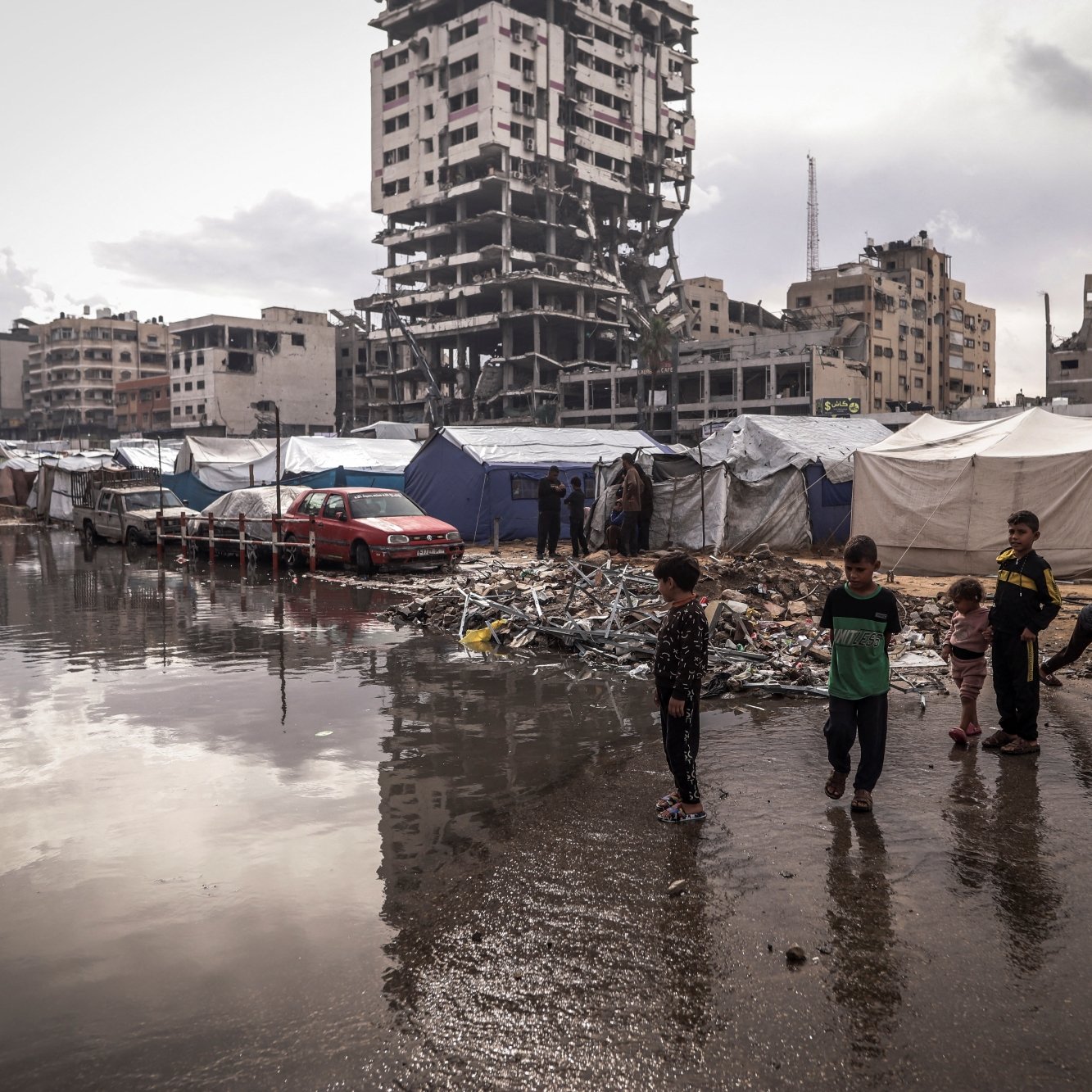 Un campo profughi con tende, bambini giocano e un edificio in rovina sullo sfondo.