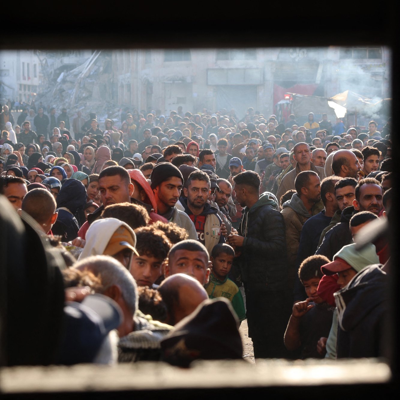 L'image montre une foule dense de personnes, probablement rassemblées dans un espace urbain. Les visages sont variés, et l'atmosphère semble chargée d'émotion et de tension. On peut voir des hommes, des femmes et des enfants, témoignant d'une situation de forte affluence, peut-être liée à un événement particulier comme une distribution d'aide ou une manifestation. Les lumières et l'arrière-plan laissent suggérer un contexte urbain avec des bâtiments visibles. L'image est encadrée par une ouverture, ce qui donne l'impression d'un regard extérieur sur ce rassemblement.