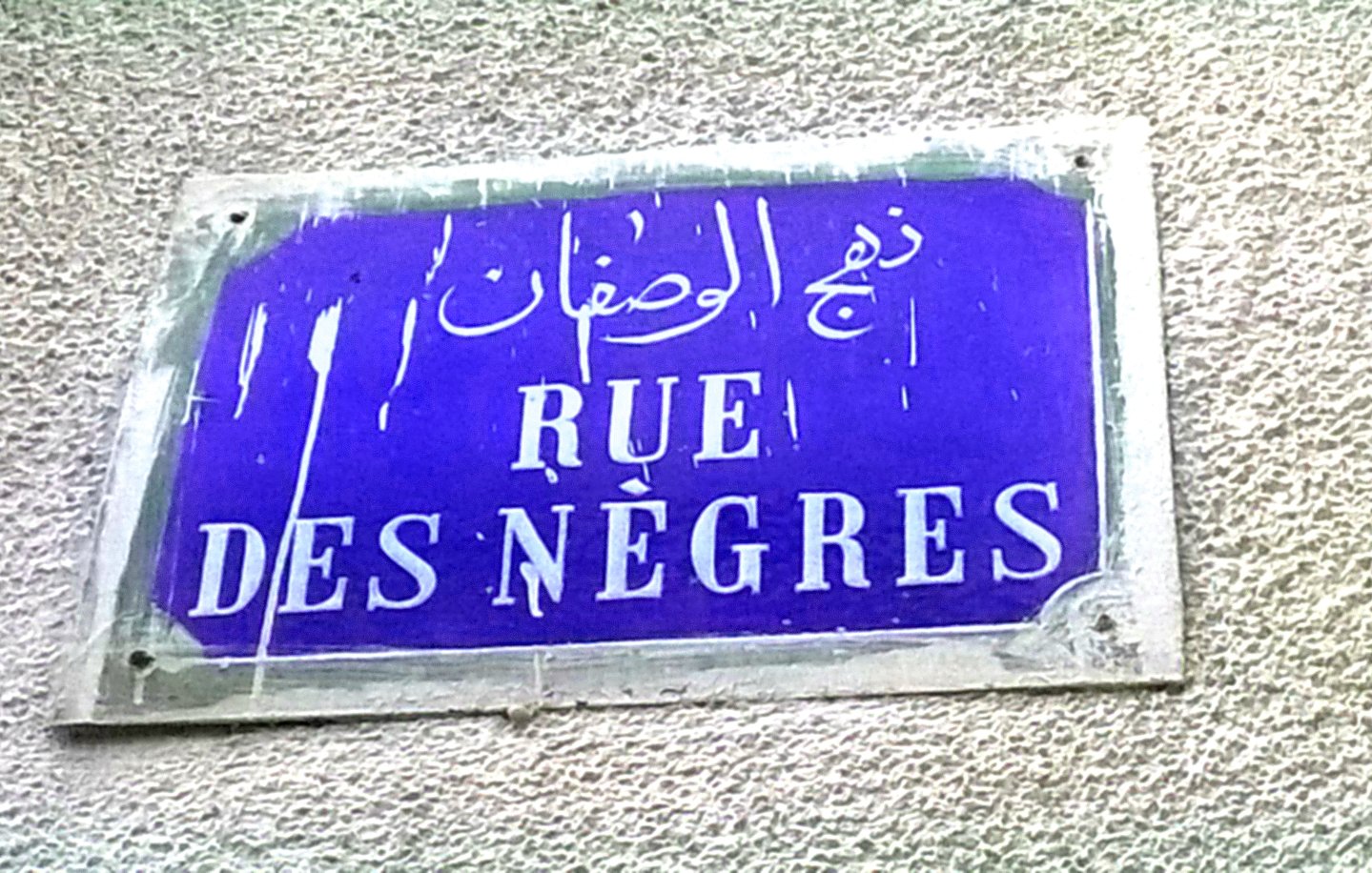 La imagen muestra una placa de calle con un fondo azul oscuro. En la placa se encuentran tres inscripciones en diferentes idiomas: árabe, francés y otro que parece ser un nombre transliterado. La parte central dice "RUE DES NÈGRES" en letras blancas. La placa parece estar un poco desgastada y sucia, lo que indica que ha estado expuesta al tiempo.