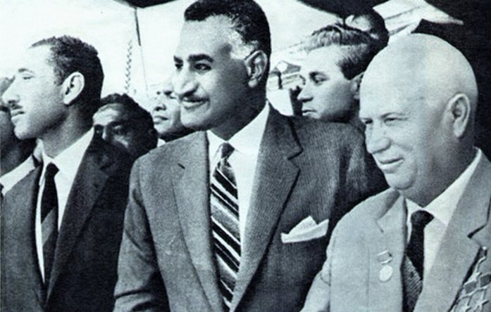 L'image montre un groupe d'hommes en costume lors d'un événement officiel. Au centre, un homme avec une moustache et une cravate rayée est entouré de deux autres individus, dont l'un se tient à sa gauche. L'ambiance semble solennelle, et ils semblent être en train de discuter ou de participer à une cérémonie. L'arrière-plan montre une foule qui semble observer l'événement.