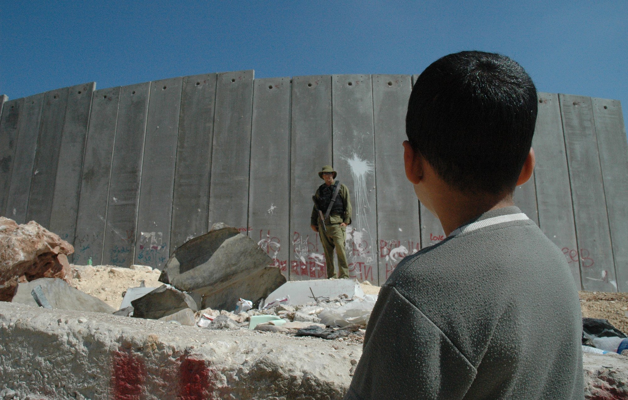 L'image montre un jeune garçon observant un soldat qui se tient devant un mur en béton. Le mur est marqué par des graffitis, suggérant un environnement de conflit ou de tension. Des débris sont dispersés au sol, et le ciel est dégagé, ce qui contraste avec la gravité de la scène. Le regard du garçon semble interrogateur ou curieux face à la situation qui l'entoure.
