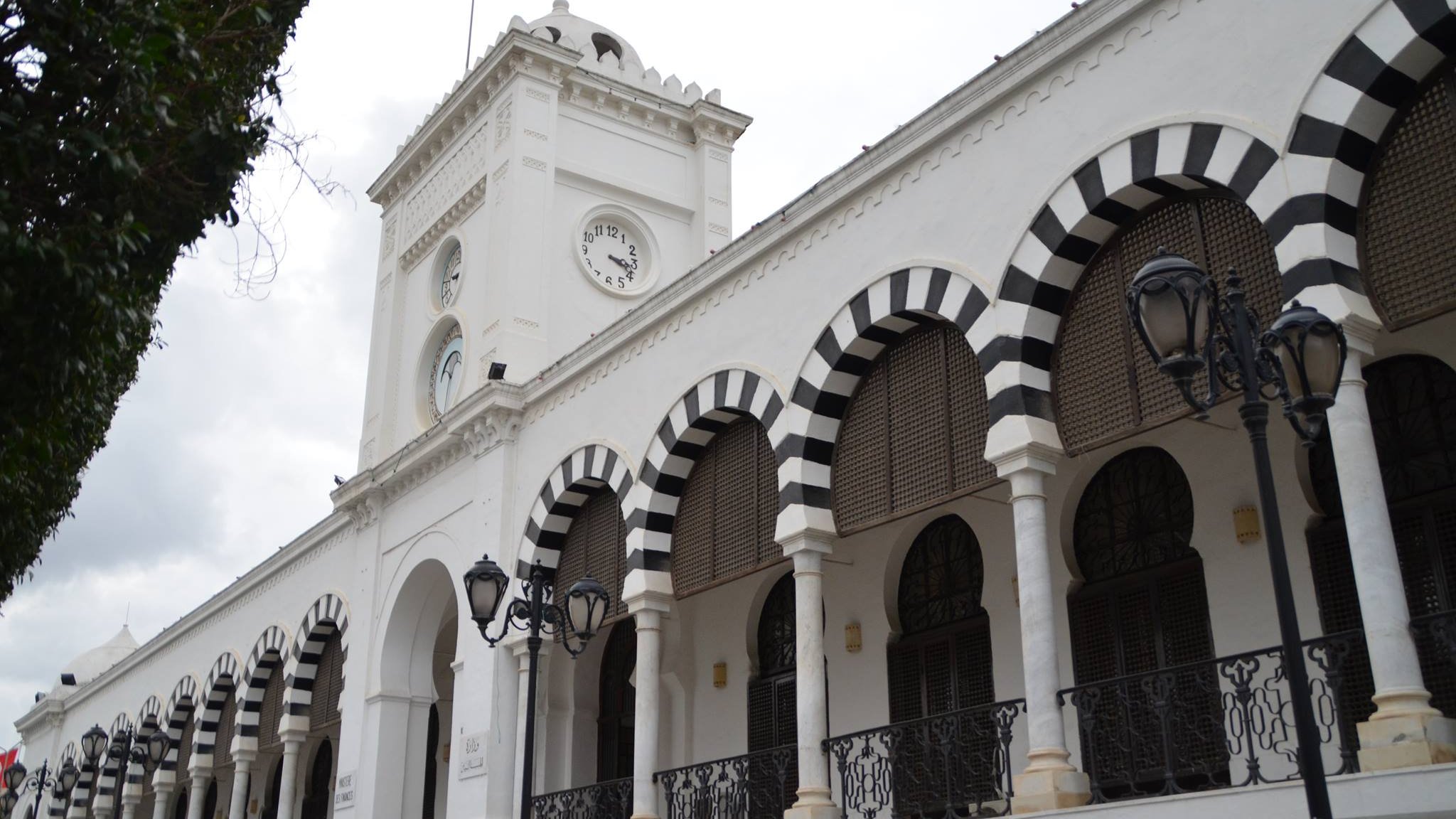 L'image montre un bâtiment architectural blanc avec des arcades noires et blanches. Il y a une horloge sur la façade et un drapeau tunisien flottant au sommet. Le ciel est nuageux, ce qui donne une ambiance légèrement sombre. Des lampadaires sont visibles le long de la structure, ajoutant une touche d'élégance à l'ensemble.