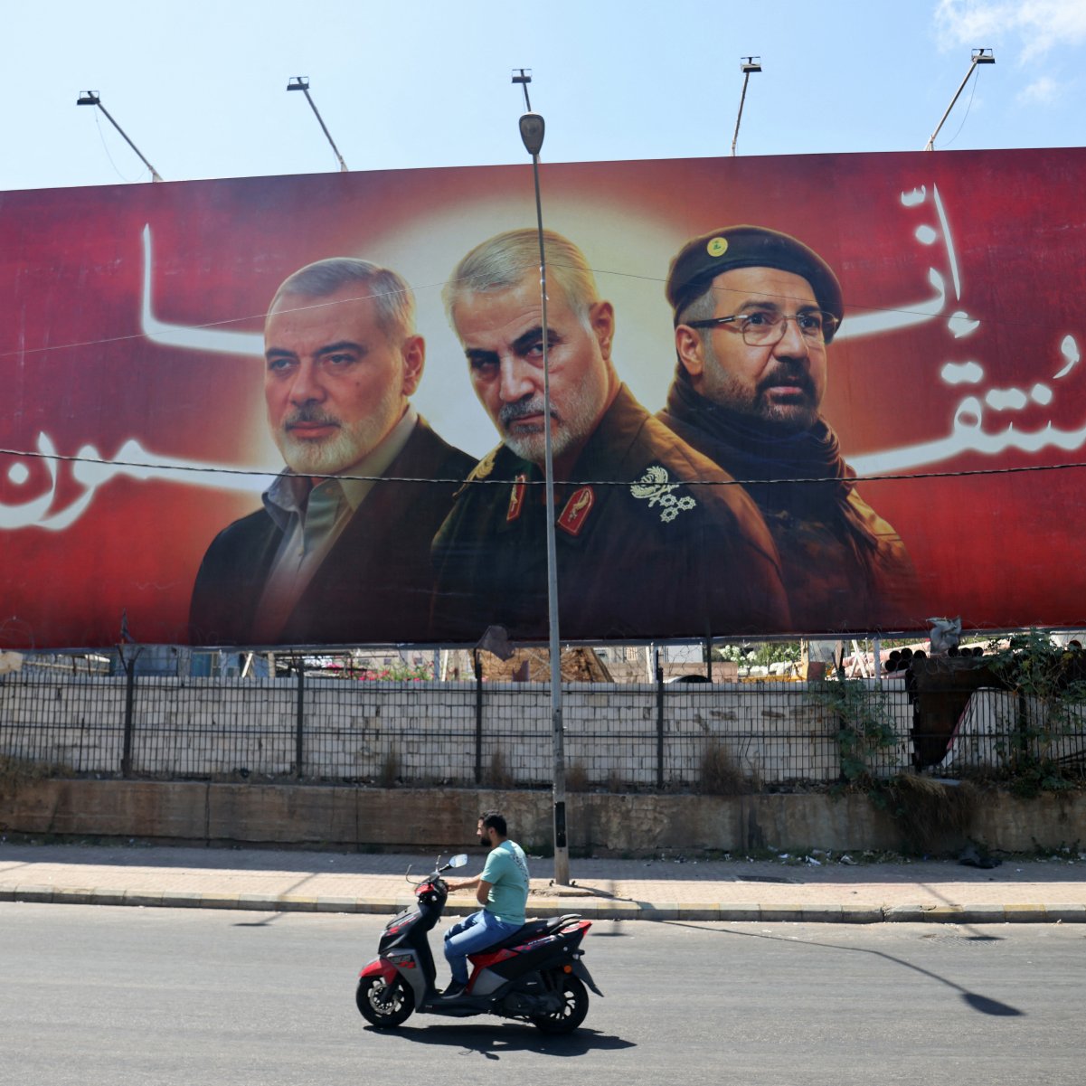 L'image montre une grande affiche publicitaire avec trois portraits d'hommes, probablement des figures militaires ou politiques, sur fond rouge. Un homme à moto passe devant cette affiche, qui semble être placée le long d'une route. L'environnement semble urbain, avec des déchets visibles au fond.