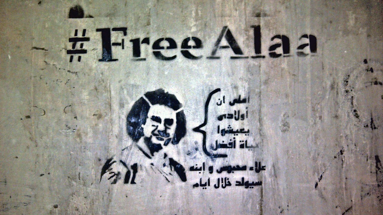 L'image présente un graffiti sur un mur. Le texte "#FreeAlaa" est inscrit en haut, accompagnant un dessin stylisé d'une personne. En dessous, il y a une citation en arabe qui semble évoquer des thèmes de liberté ou de résistance. L'ensemble donne une impression de soutien à une cause ou un individu en particulier, en soulignant la lutte pour la liberté d'expression.