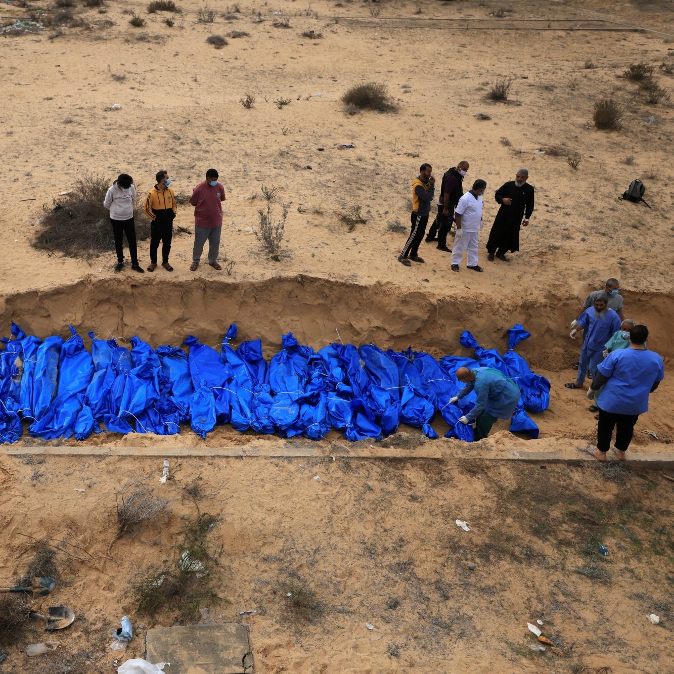 تصویر، گودالی در بیابان با تعدادی کیسه آبی رنگ و افرادی در حال مشاهده یا کار در اطراف آن است.