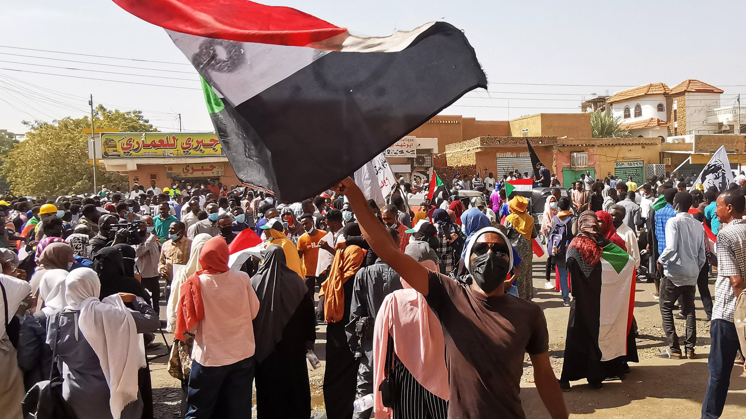 L'image montre une grande foule réunie dans une manifestation, avec des personnes brandissant des drapeaux. Au premier plan, un individu tient un drapeau soudanais, et de nombreuses autres personnes portent des masques. L'atmosphère semble énergique et mobilisée, avec des pancartes et des expressions de détermination. En arrière-plan, on peut apercevoir des bâtiments et des enseignes, indiquant un cadre urbain.