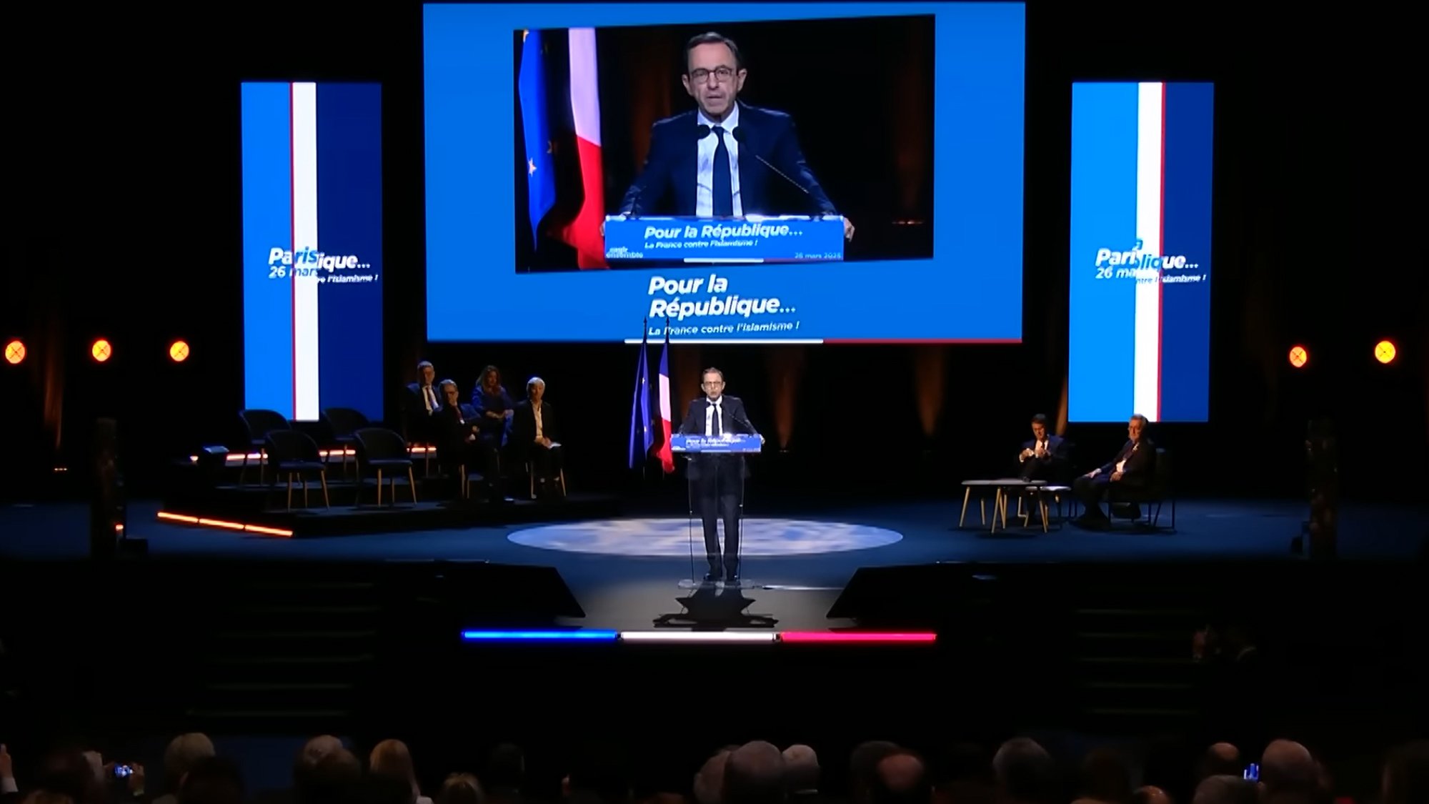 Un homme parle sur scène, avec des drapeaux français en arrière-plan. 