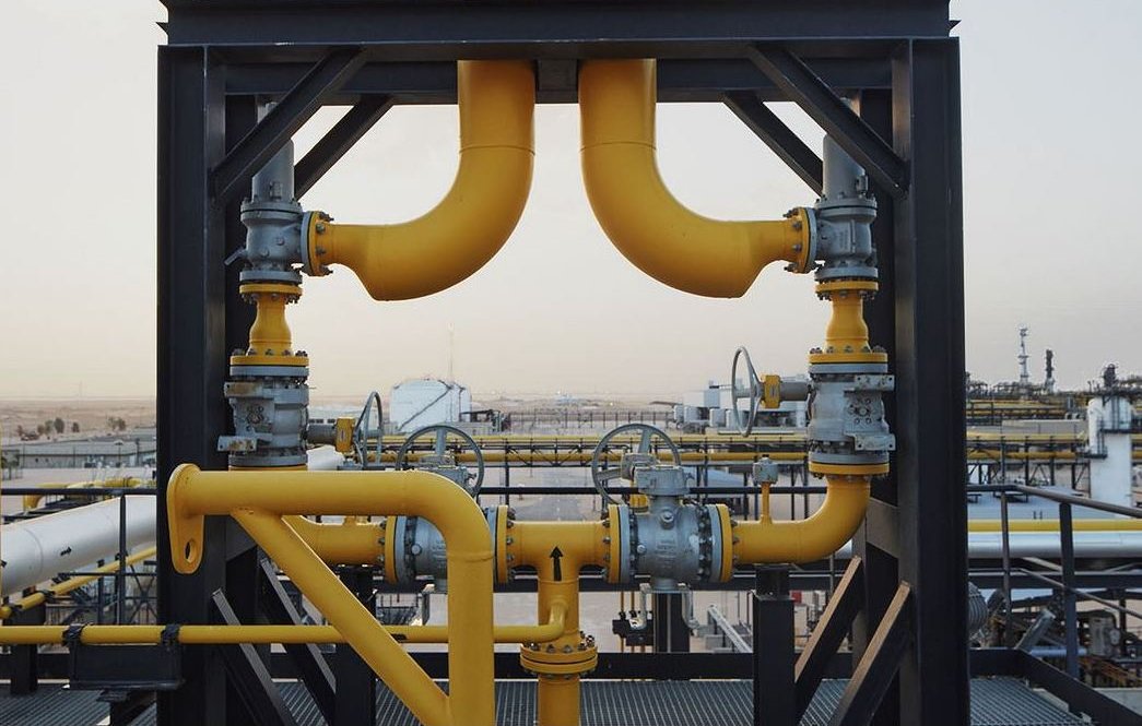 La imagen muestra una estructura industrial, probablemente de una planta de procesamiento de petróleo o gas. Hay varias tuberías de color amarillo que se conectan entre sí, junto con válvulas y otros elementos metálicos. El fondo sugiere un paisaje industrial con tanques y equipos, lo que indica un entorno de trabajo relacionado con la energía o la petroquímica. La escena parece ser al aire libre, bajo un cielo claro.