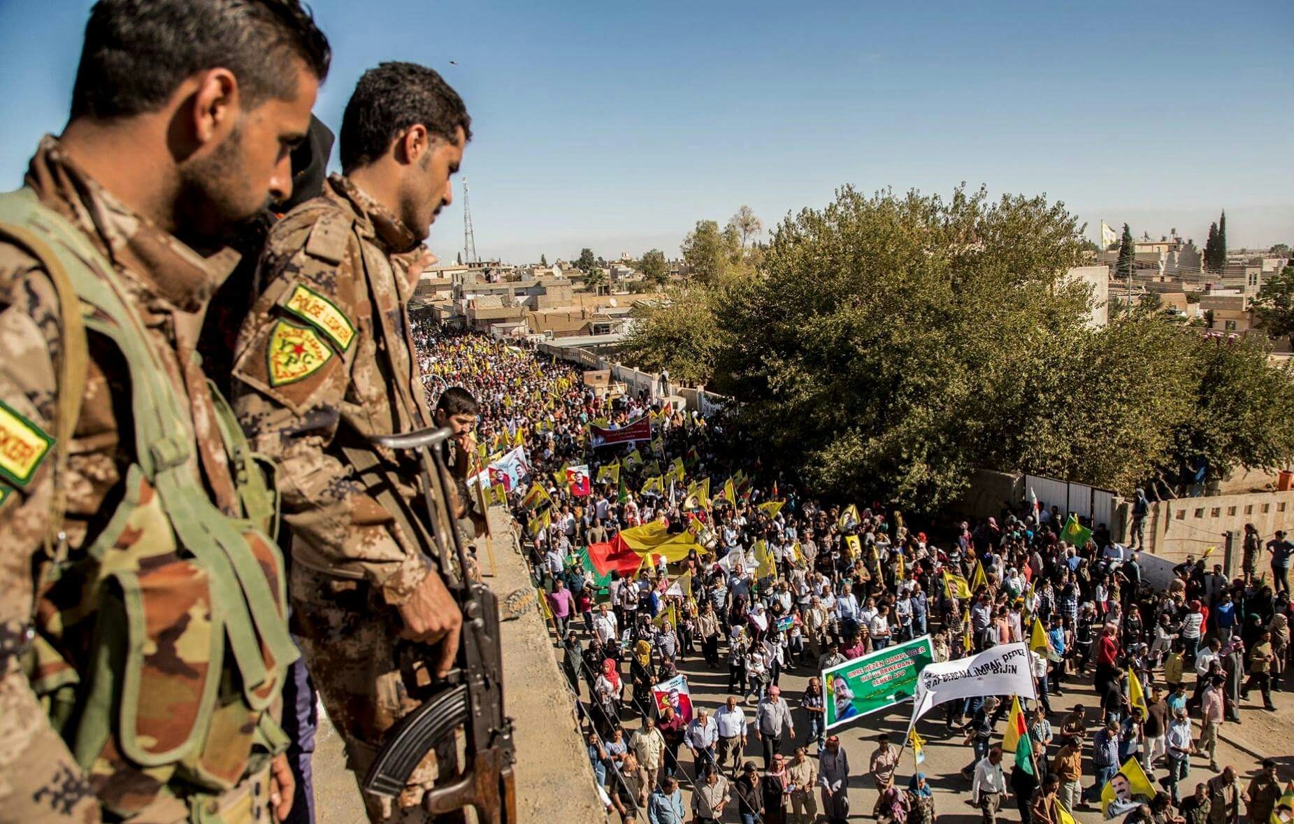 The image depicts a large crowd participating in a demonstration or march. In the foreground, two armed soldiers in military uniforms are positioned on a higher vantage point, observing the event. Below them, a diverse group of people holds various flags and banners, indicating that the gathering is likely a political or social protest. The scene is set in an outdoor urban environment, with trees and buildings visible in the background. The atmosphere appears to be one of engagement and collective action.
