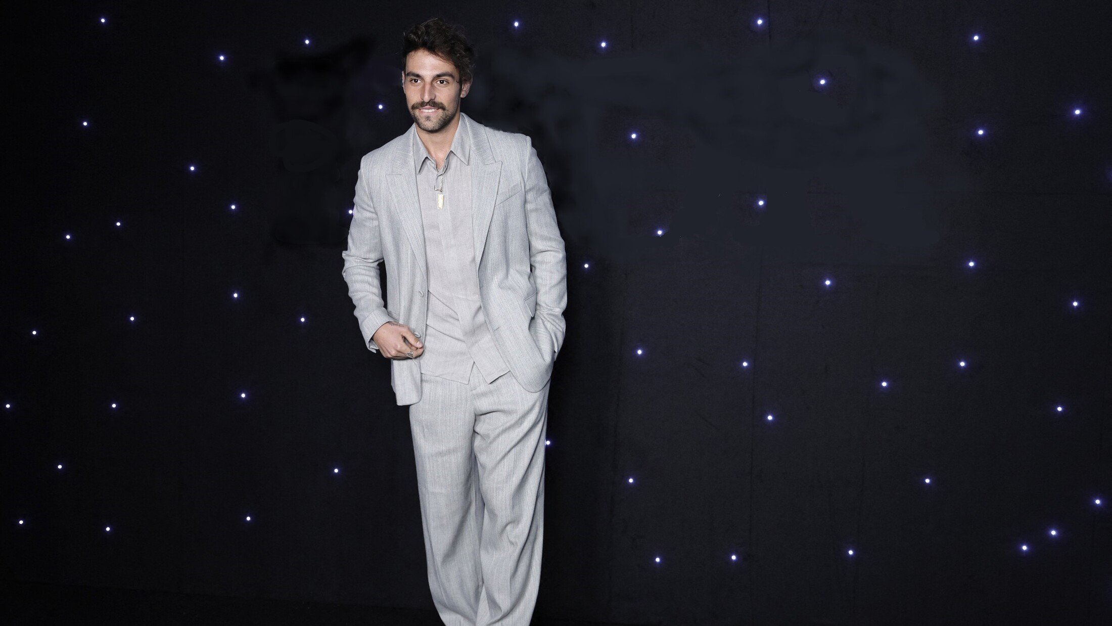 L'image montre un homme portant un costume léger à rayures, composé d'une veste et d'un pantalon ample. Il a une moustache et un sourire, et se tient debout avec une pose décontractée. L'arrière-plan est sombre, parsemé de petites lumières blanches qui ajoutent un effet élégant à la scène.