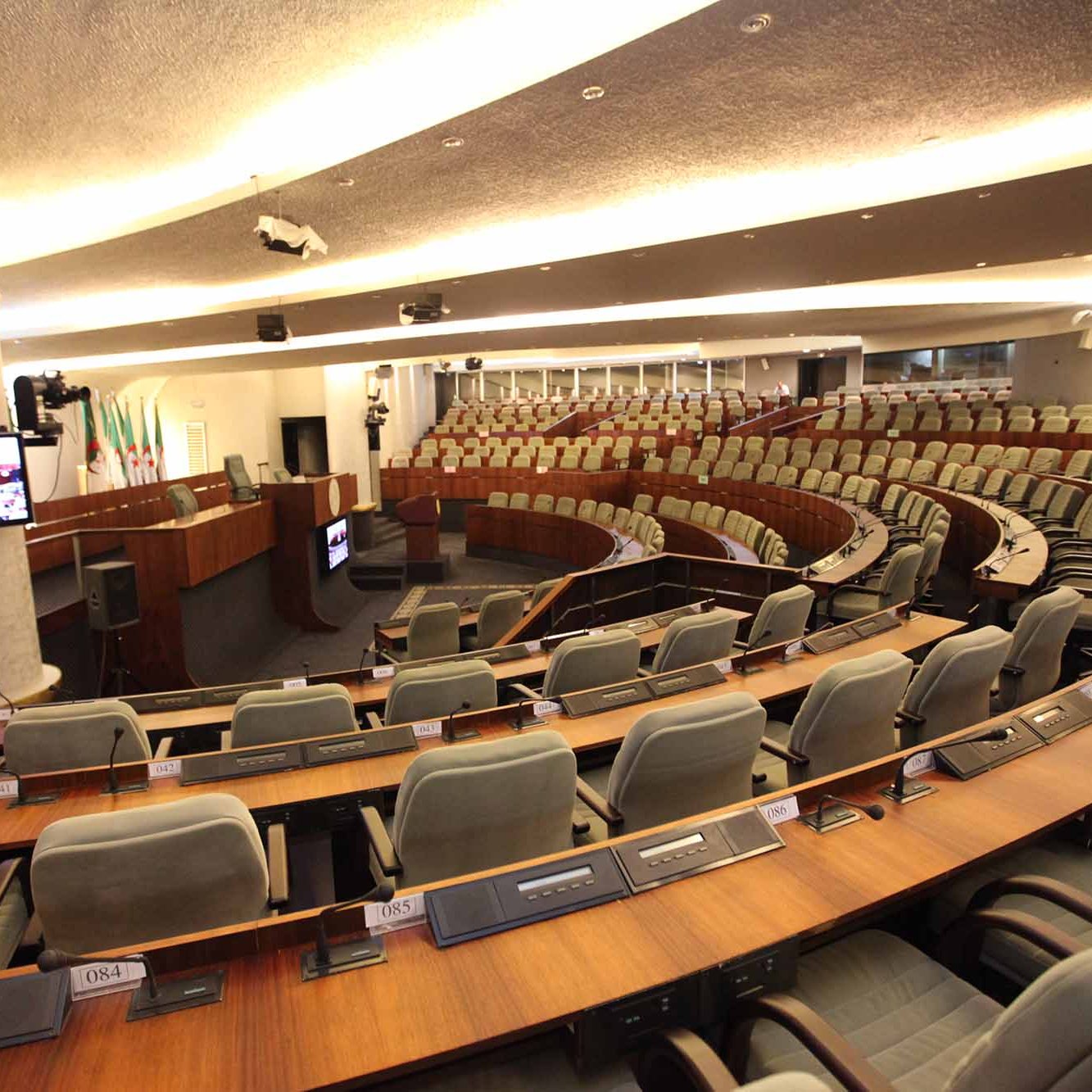 L'image représente une salle de réunion ou un hémicycle, typique des assemblées législatives. On peut voir des sièges disposés en amphithéâtre, avec des rangées de fauteuils gris. À l'avant, il y a une estrade avec un bureau et des équipements audiovisuels. La décoration est moderne et sobre, montrant un environnement formel adapté aux discussions et aux débats. Des écrans sont également présents pour fournir des informations aux membres présents.