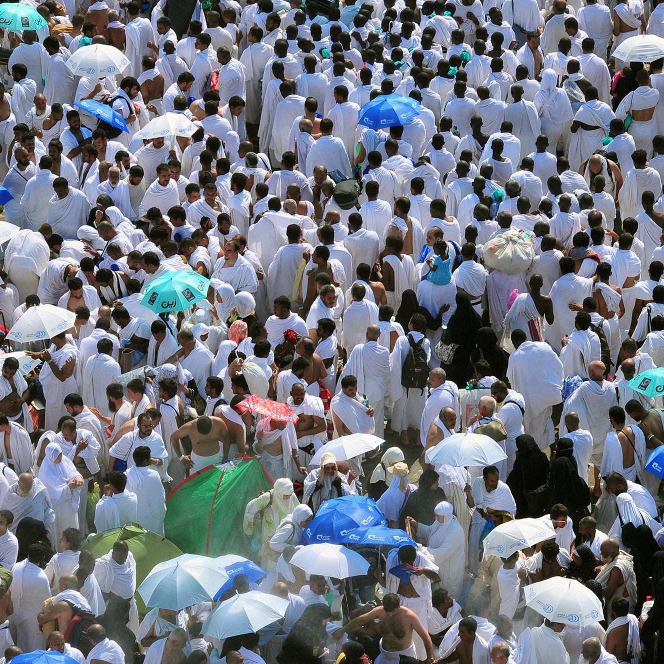 L'image montre une grande foule de personnes vêtues de vêtements blancs, probablement en train de participer à un pèlerinage ou à un événement religieux. De nombreux participants arborent des parapluies colorés, qui offrent de l'ombre dans un environnement ensoleillé. L'atmosphère semble très animée, avec un grand nombre de personnes rassemblées, ce qui reflète un moment de rassemblement spirituel.