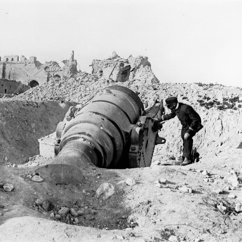 L'image montre un homme travaillant près d'un grand canon ou d'une pièce d'artillerie en position, probablement dans un environnement de guerre. À l'arrière-plan, on peut voir des bâtiments en ruine, ce qui suggère que la scène se déroule dans une zone de conflit, possiblement après un bombardement. Le paysage est aride et rempli de débris, ce qui accentue l'atmosphère de destruction. L'homme semble concentré sur son travail, illustrant les efforts militaires dans un contexte difficile.