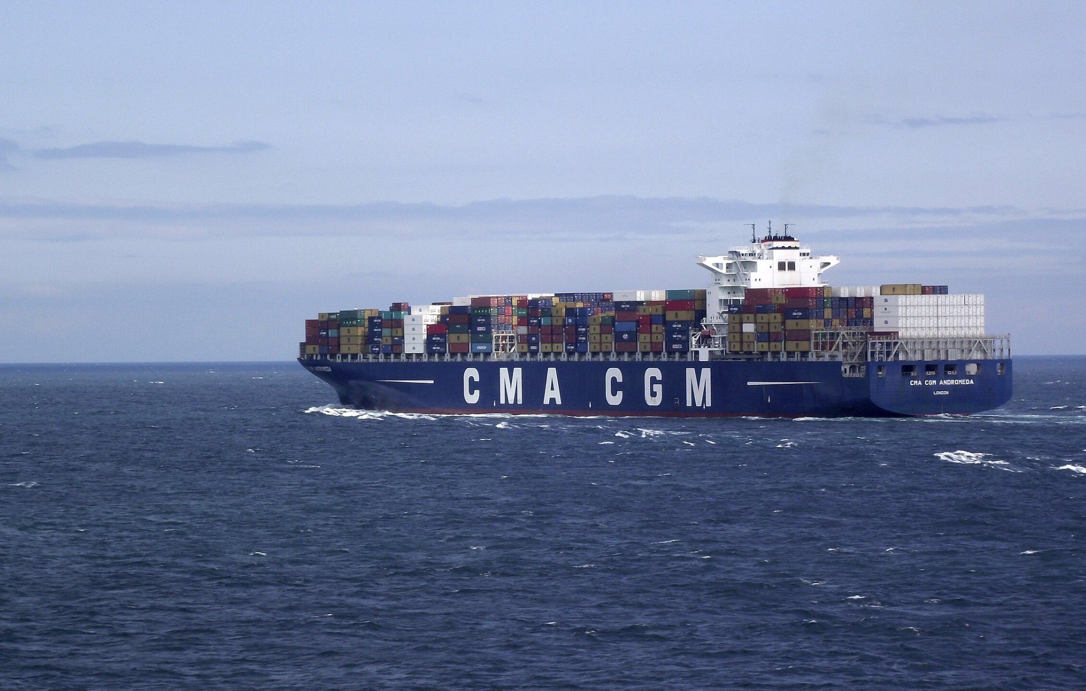 L'image montre un grand porte-conteneurs naviguant sur une mer calme. Le navire, qui porte les lettres "CMA CGM" sur son flanc, est couvert de conteneurs multicolores empilés sur le pont. Le ciel est légèrement nuageux, avec des teintes de bleu, et on peut apercevoir des vagues sur l'eau. L'ensemble dégage une impression de commerce maritime et de transport international.