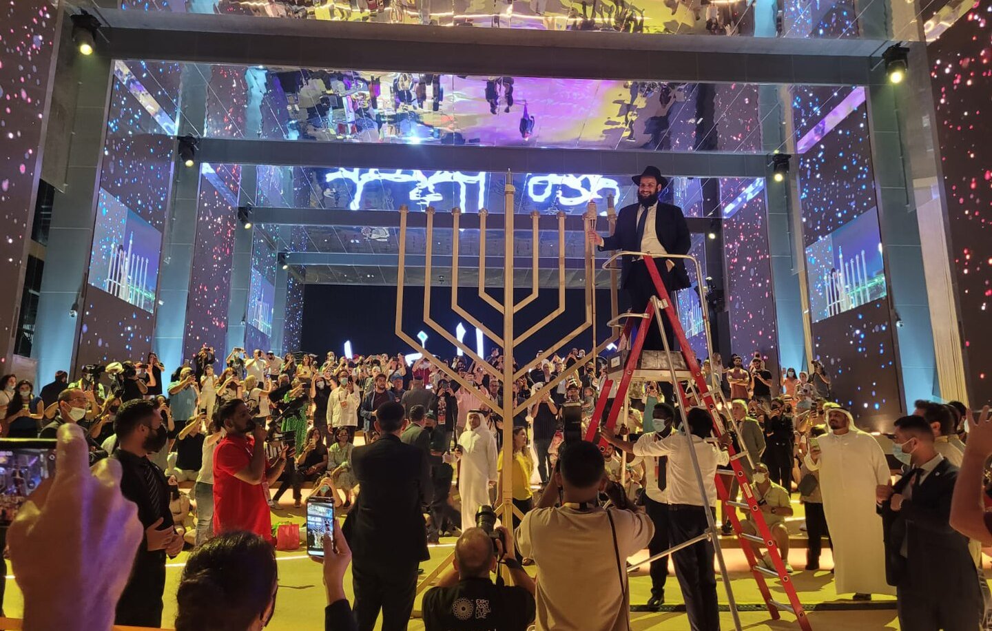 L'image montre une scène festive dans une grande salle. Au centre, une grande menorah (un candélabre à sept branches) est mise en avant. Un homme en costume est sur une échelle, probablement en train d'allumer une bougie. La salle est remplie de personnes, certaines prenant des photos, tandis que d'autres regardent attentivement. Les murs sont décorés avec des éclairages colorés et des projections, créant une atmosphère vibrante et festive. L'ensemble de l'événement semble célébrer une occasion spéciale, probablement liée à la culture ou à la tradition juive.