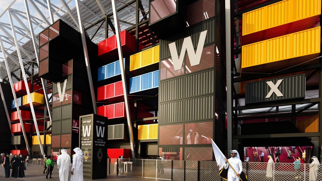 La imagen muestra una estructura arquitectónica moderna y colorida, posiblemente un estadio o un evento relacionado con el deporte. El diseño utiliza contenedores de carga en varios colores, que están dispuestos de manera creativa. En primer plano, se pueden ver algunas personas, algunas vestidas con trajes tradicionales, que parecen estar observando y visitando el lugar. El ambiente es vibrante y contemporáneo, destacando la innovación en la construcción.