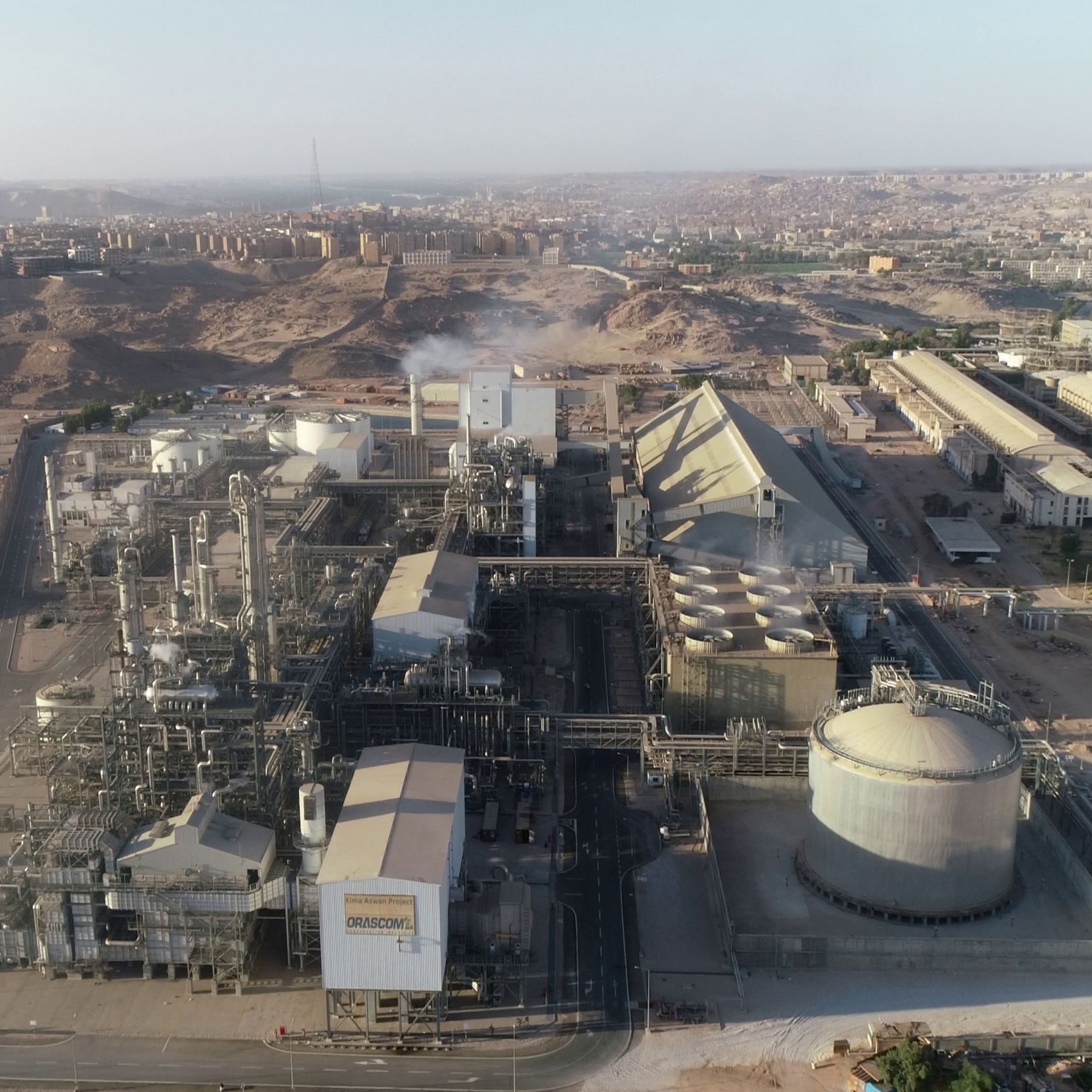 L'image présente une vue aérienne d'un complexe industriel. On peut observer plusieurs bâtiments de grande taille, comprenant des structures cylindriques et pyramidales. La zone environnante semble aride avec des collines et des bâtiments à l'arrière-plan. Des tuyaux et des infrastructures industrielles sont visibles, indiquant une activité de production. On discerne également de la vapeur s'échappant d'une des installations, suggérant un processus en cours. L'ensemble dénote un environnement industriel dans une région désertique.