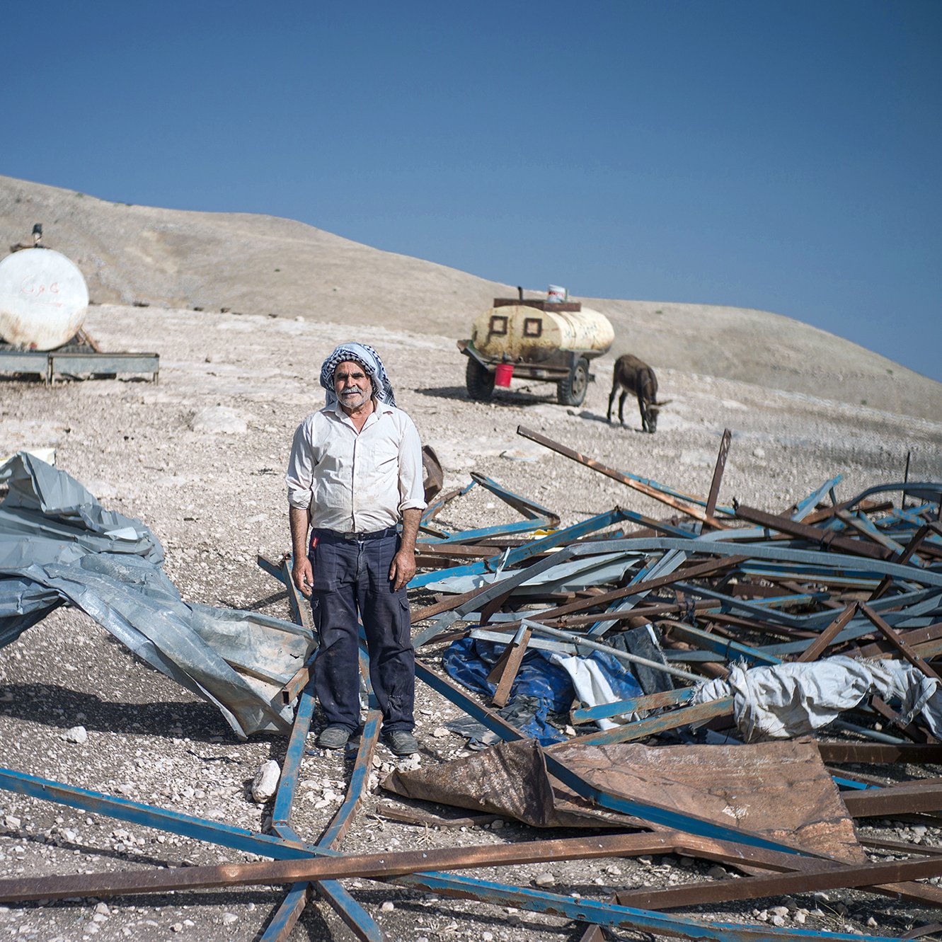 الصورة تظهر رجلًا standing في منطقة قاحلة، محاطًا بحطام وقطع من المعدن والمواد الأخرى. يبدو أن الرجل يرتدي ملابس تقليدية، وهناك خلفه مركبات ووحدات تخزين، كما تبدو الأرض غير مستوية وتنتشر فيها الصخور والحطام. السماء واضحة وزرقاء، مما يدل على يوم مشمس.