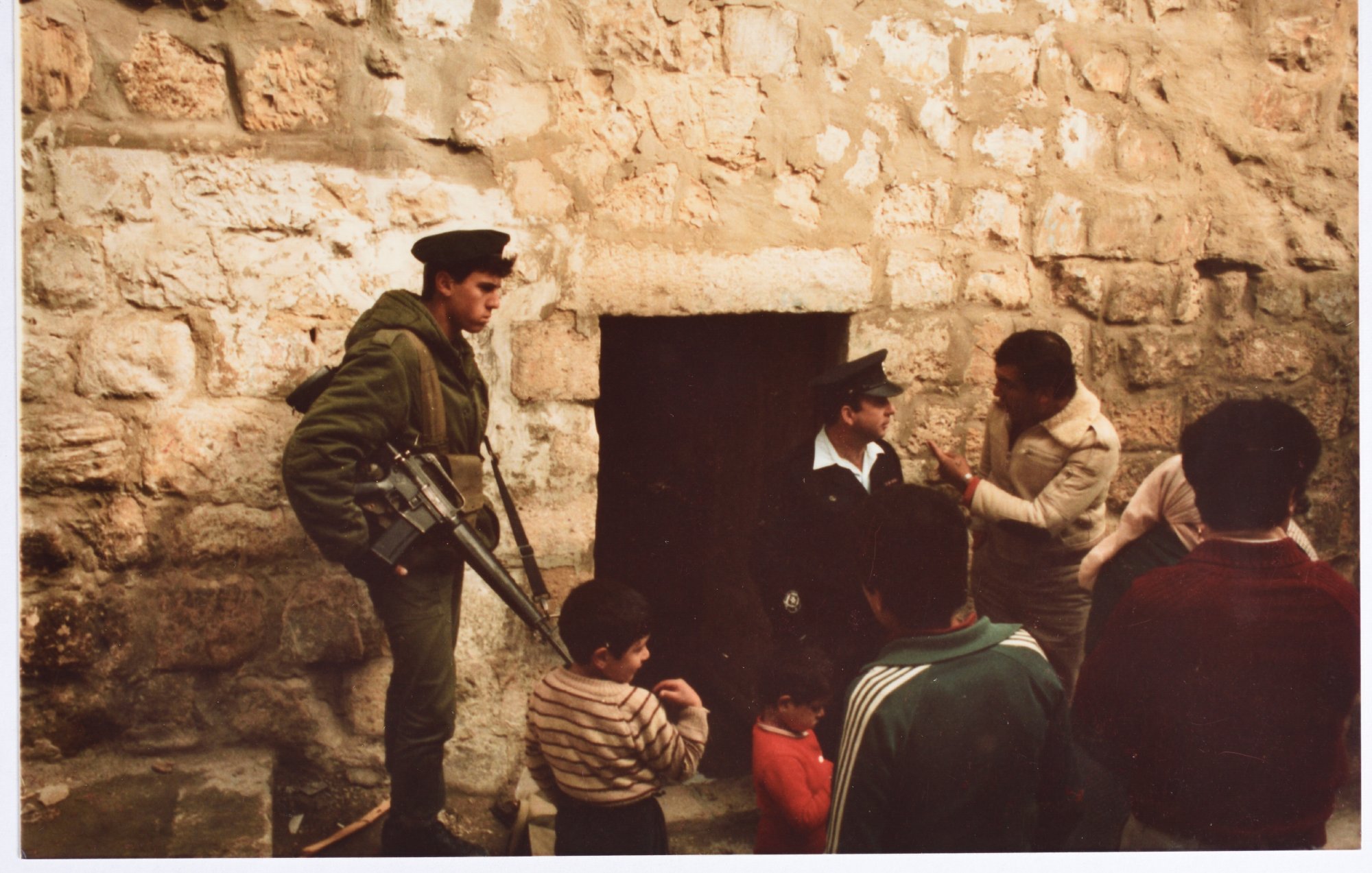 L'image montre un groupe de personnes près d'une porte en pierre. À gauche, un soldat armé en uniforme vert se tient debout, gardant l'entrée. Deux hommes adultes discutent, tandis qu'un enfant et un autre homme semblent observer la scène. L'environnement est marqué par des murs de pierre brute, suggérant un endroit ancien ou historique. L'atmosphère paraît chargée, et les expressions des personnes indiquent une interaction sérieuse.