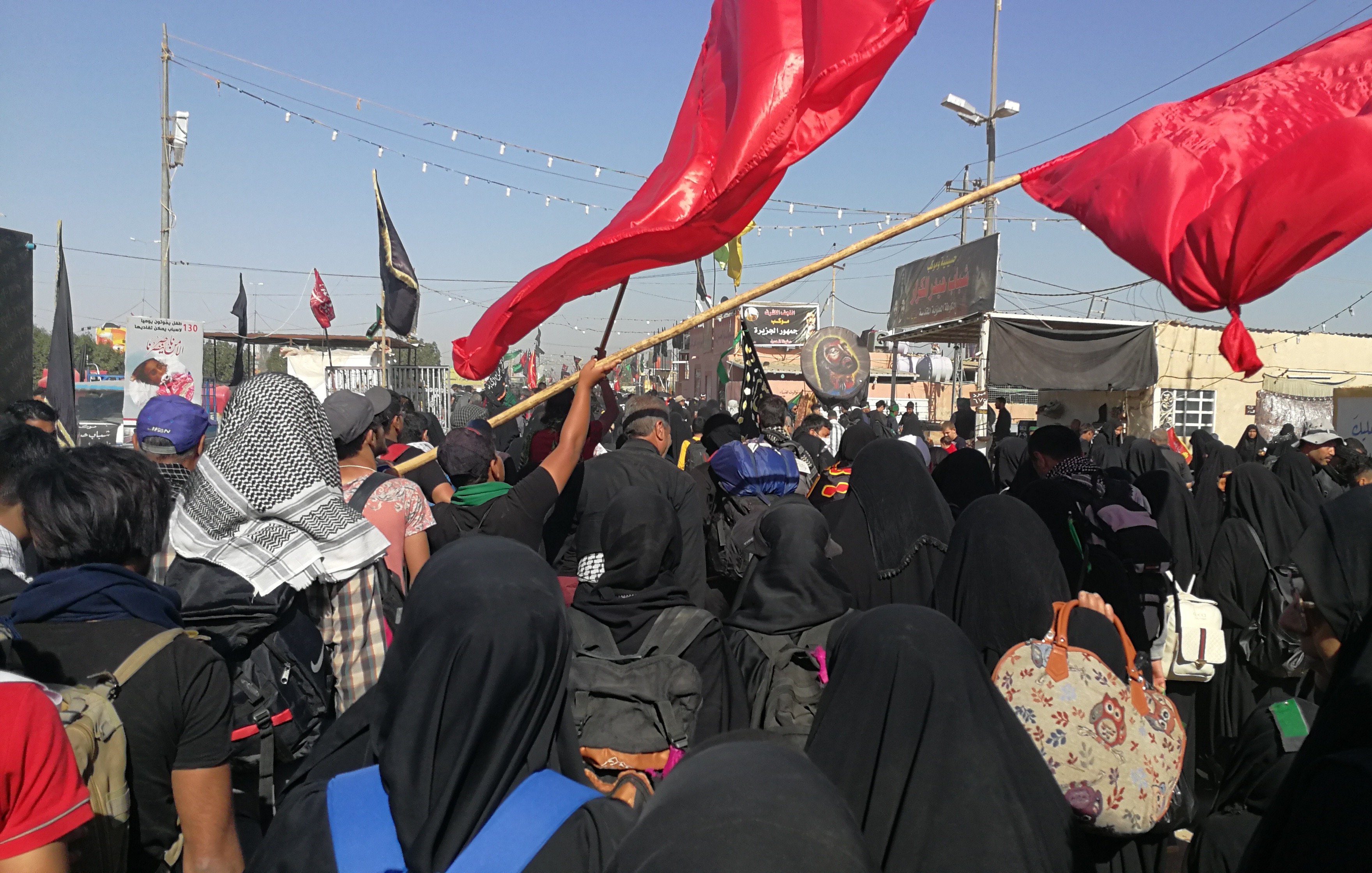 تظهر في الصورة مجموعة كبيرة من الأشخاص، معظمهم يرتدون الملابس السوداء، مما يشير إلى أنهم ربما يشاركون في مناسبة دينية. يحمل بعضهم أعلامًا حمراء، وهناك لافتات وأعلام أخرى في الخلفية. الأجواء تظهر حماسًا وجمعًا كبيرًا من الناس، مما يعكس أهمية الحدث الذي يشتركون فيه. السماء صافية وأشعة الشمس تضيء المكان.