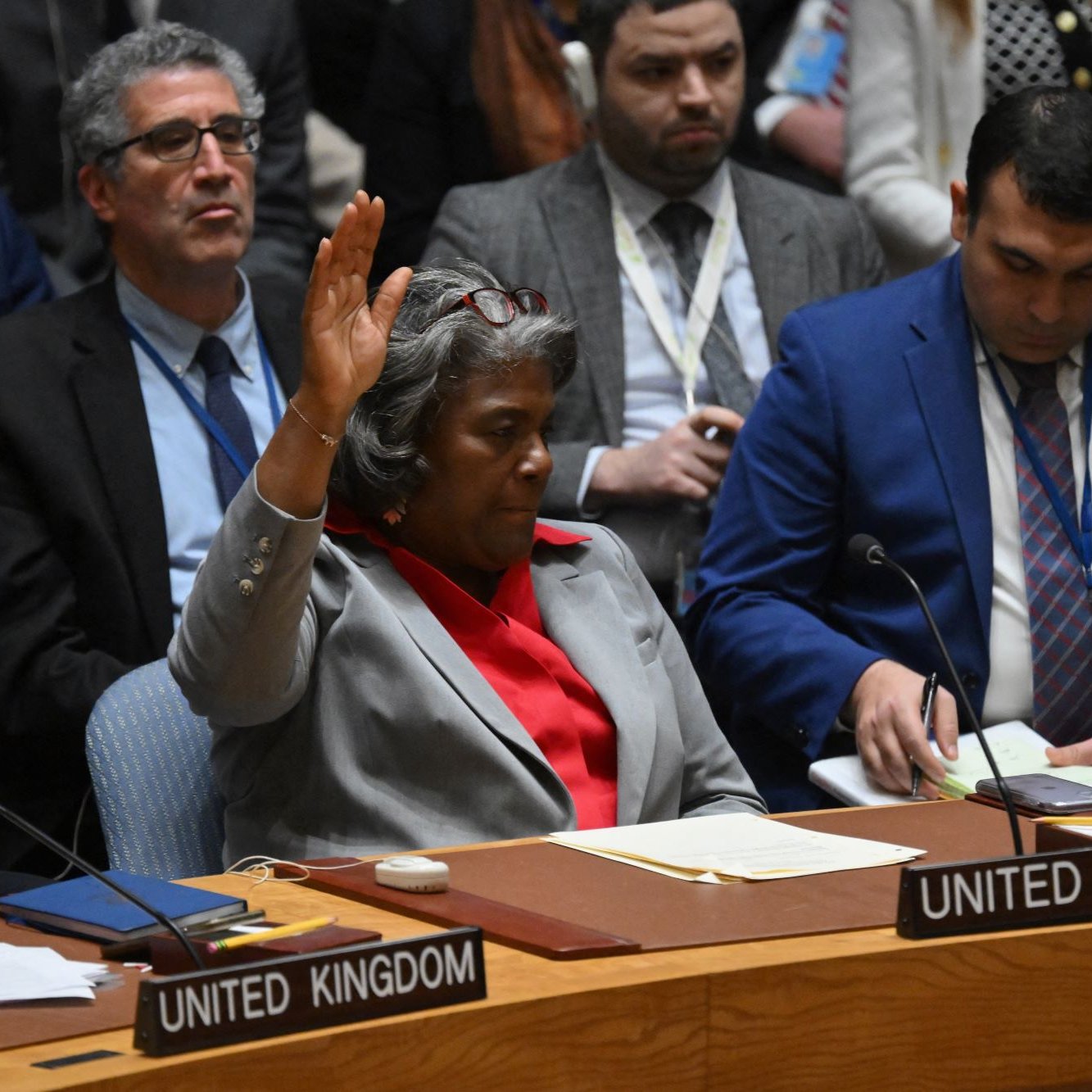 L'image montre une réunion au Conseil de sécurité des Nations Unies. Une femme, manifestement représentant les États-Unis, est assise à une table, levant la main pour prendre la parole ou voter. À côté d'elle, on aperçoit d'autres délégués de différents pays, tous attentifs. L'atmosphère semble sérieuse et concentrée, typique des discussions diplomatiques importantes.