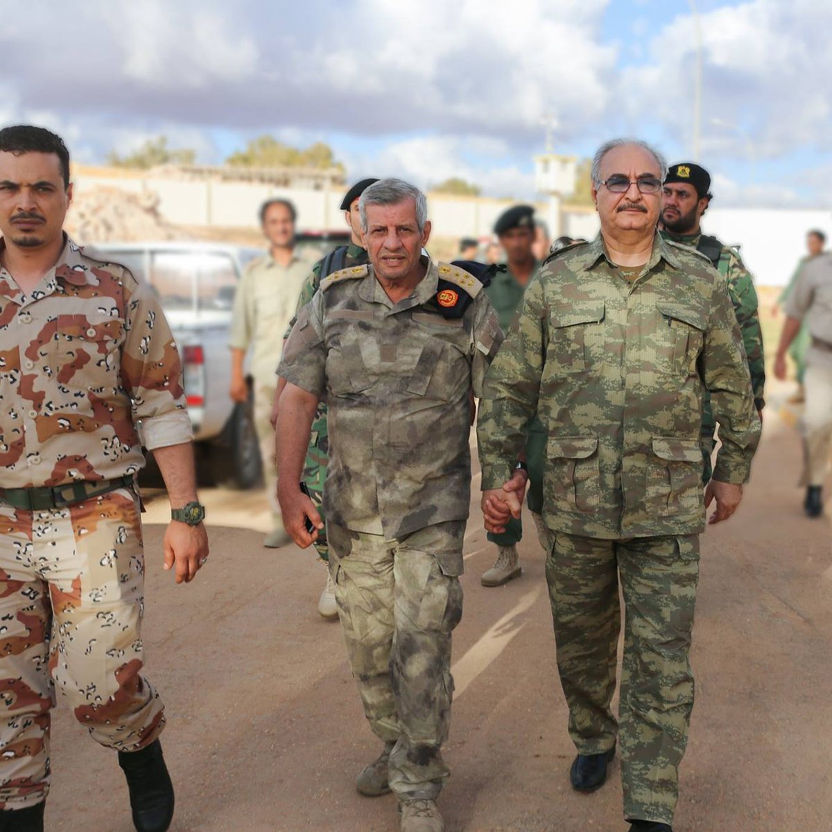 The image depicts a group of military personnel walking together. Two men in the foreground, wearing military uniforms, are holding hands and appear to be engaged in conversation. One is in a camouflage uniform, while the other has a distinctive pattern. They are flanked by soldiers in various military attire in the background. The setting appears to be an outdoor area, possibly a military base, with vehicles and other personnel visible. The sky shows some clouds, indicating daytime.