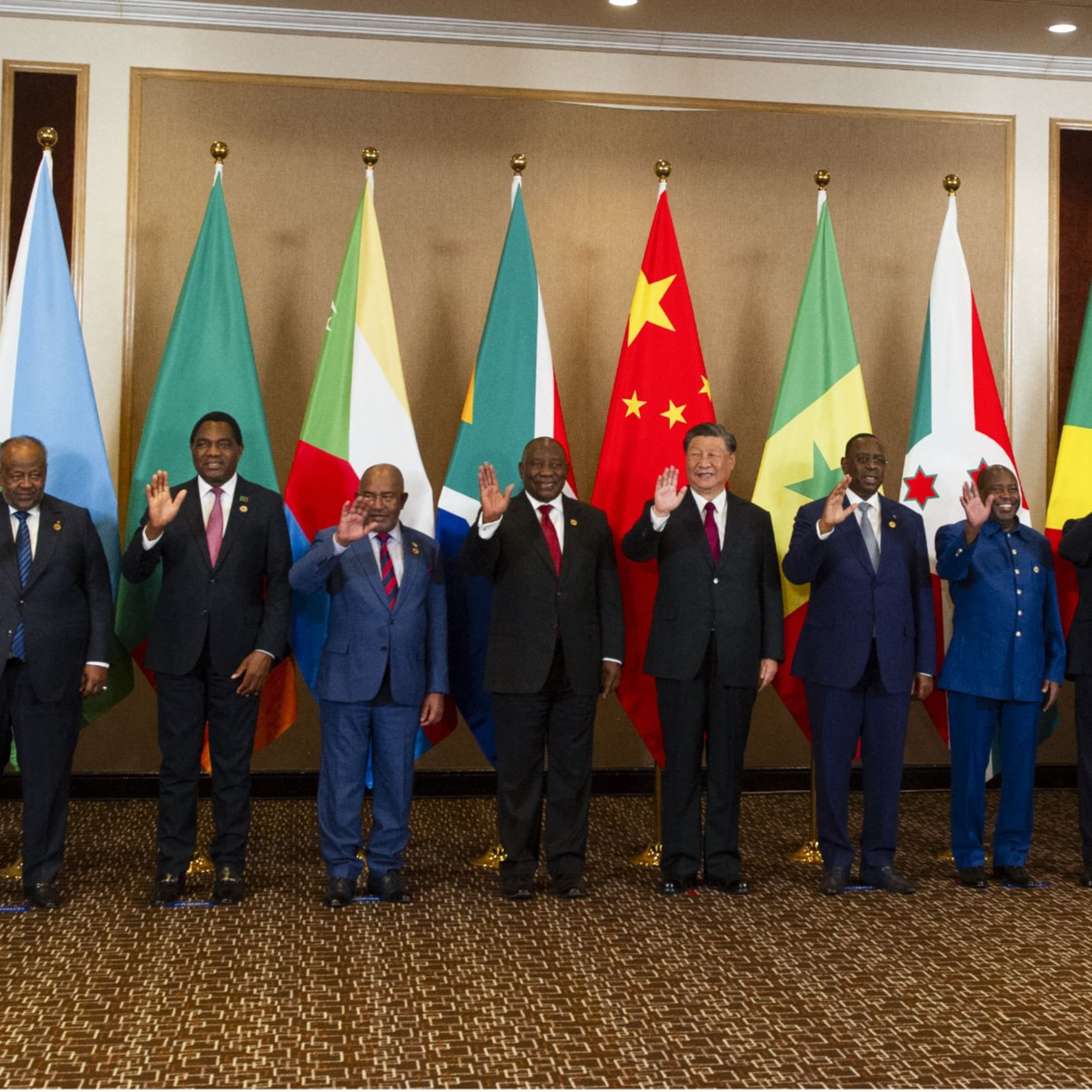The image features a group of politicians standing together in a formal setting. They are arranged in a line, with each person smiling and waving. Behind them, there are several national flags, including those of various countries such as China and several African nations, indicating an international or diplomatic event. The individuals appear to represent different countries or organizations, reflecting a multicultural gathering. The setting has an elegant interior, suggesting a significant and official occasion.