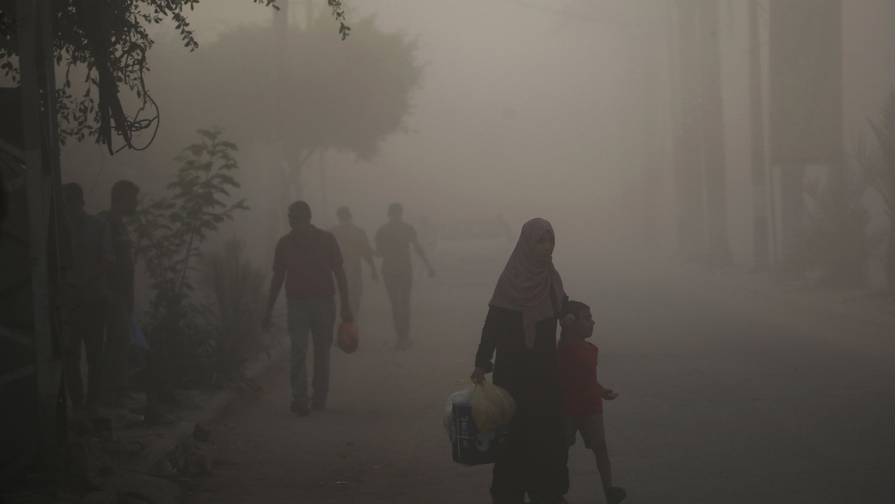 L'immagine mostra una scena di una strada avvolta nella nebbia o nella polvere. In primo piano, una donna con un velo scuro tiene la mano di un bambino, mentre camminano insieme. Sullo sfondo, si possono vedere altre persone che camminano, ma sono poco visibili a causa della foschia. Gli alberi e alcune piante ai lati della strada creano un'atmosfera un po' opprimente. La luce sembra attenuata, dando un'impressione di desolazione.