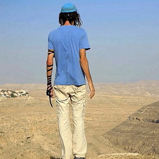 Cette image montre une personne de dos, se tenant au bord d'une falaise. Elle porte un t-shirt bleu et des pantalons clairs, ainsi qu'une kippa sur la tête. Son bras gauche est vêtu d'un bandage. Le paysage environnant est désertique, avec des collines et une vallée visible à l'arrière-plan. On peut apercevoir quelques maisons au loin, suggérant une présence humaine dans cette vaste étendue. L'atmosphère est calme, avec un ciel clair.