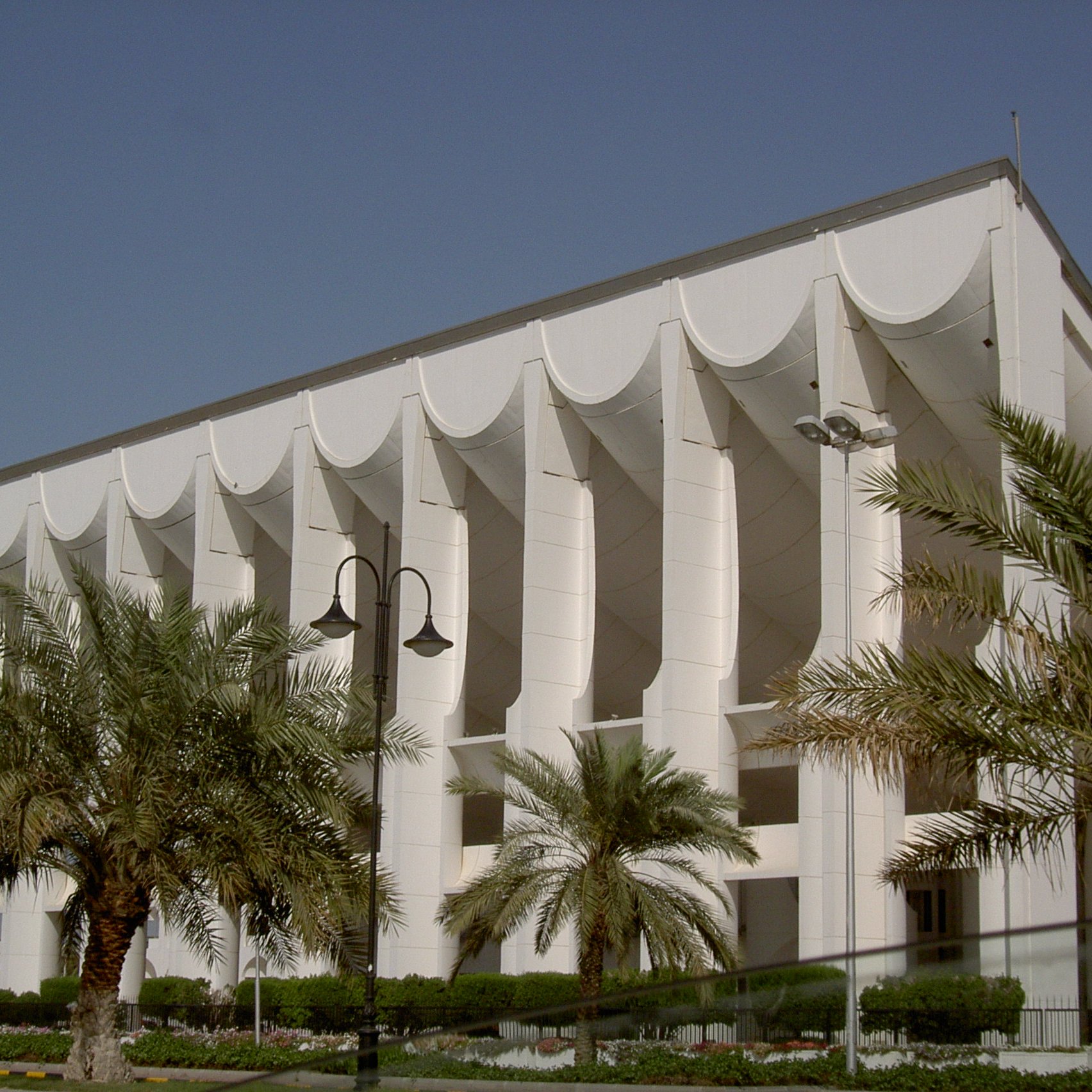 L'image montre un bâtiment moderne avec une architecture distinctive. La façade est principalement blanche, ornée de structures en forme de vagues ou de voiles sur le haut. Des colonnes soutiennent la structure, ajoutant une touche de sophistication. On peut également observer des palmiers plantés devant le bâtiment, ce qui donne une atmosphère tropicale et accueillante. Le ciel est clair et bleu, accentuant la luminosité de l'ensemble.