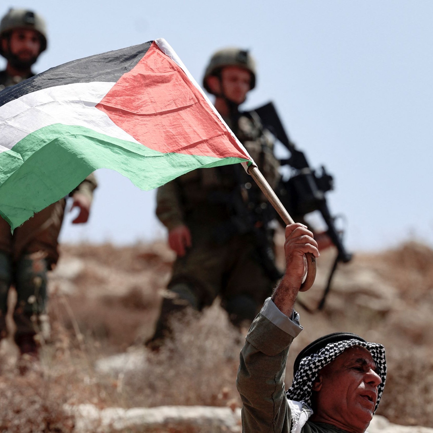 L'image montre un homme brandissant fièrement un drapeau palestinien. Il semble être en première ligne, tandis que des soldats en arrière-plan, probablement des soldats israéliens, sont visibles. Le paysage est sec et rocailleux, suggérant un environnement rural ou semi-aride. L'atmosphère semble tendue en raison de la présence militaire et du geste de protestation de l'homme.