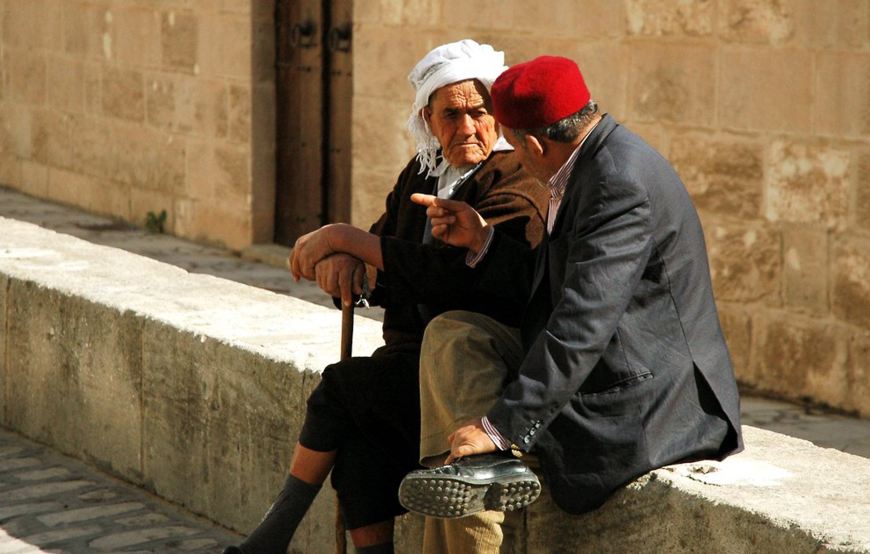 Deux hommes âgés discutent assis sur un mur en pierre, l'un portant un fez rouge.