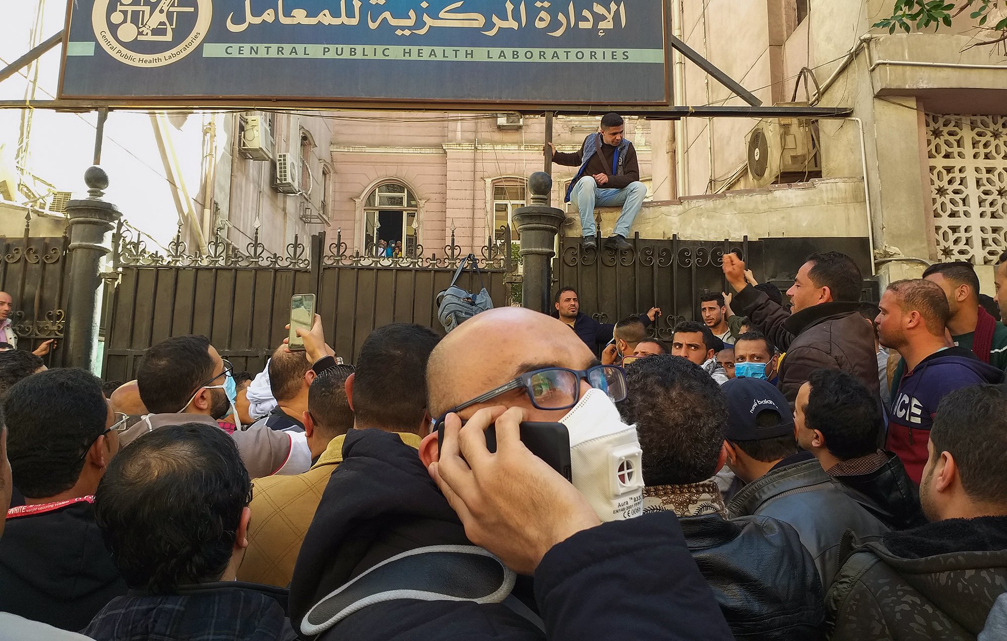 L'image montre une foule rassemblée devant un bâtiment marqué par une enseigne en arabe et en anglais, indiquant qu'il s'agit des laboratoires de santé publique. Des hommes sont visibles, certains portant des masques, tandis qu'un individu est assis sur une barrière. L'atmosphère semble tendue ou agitée, et les personnes semblent discuter ou crier. Le cadre urbain suggère un contexte de mobilisation ou de manifestation.