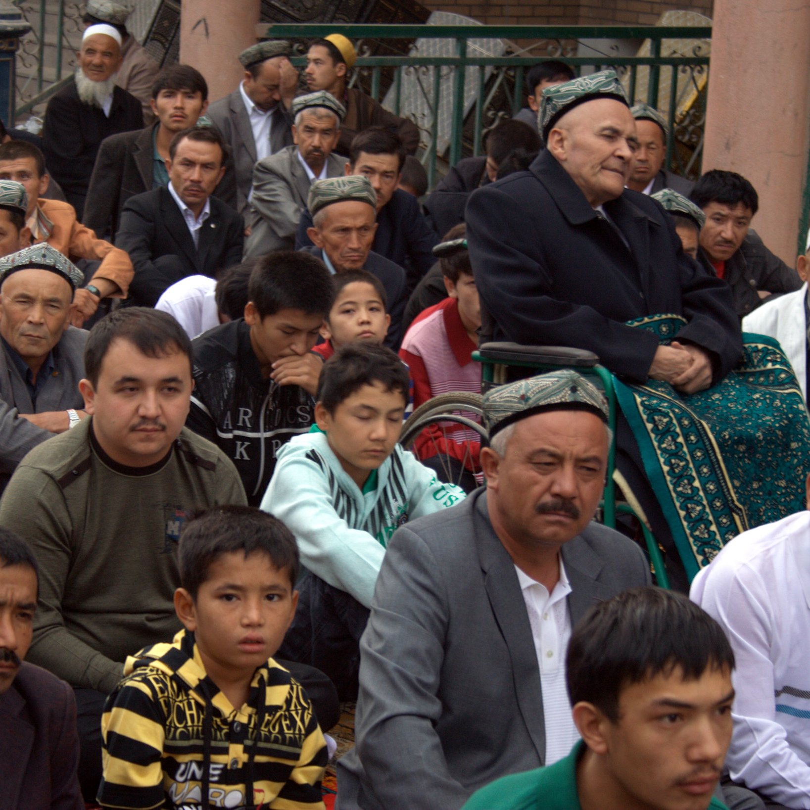 L'image montre un groupe de personnes assises, probablement lors d'un rassemblement ou d'une prière. On voit des hommes de différentes générations, certains portant des chapeaux traditionnels. Au centre, un homme âgé est assis sur une chaise, entouré de jeunes et d'adultes qui semblent attentifs. L'ambiance est sérieuse et respectueuse, reflétant un moment important pour la communauté.